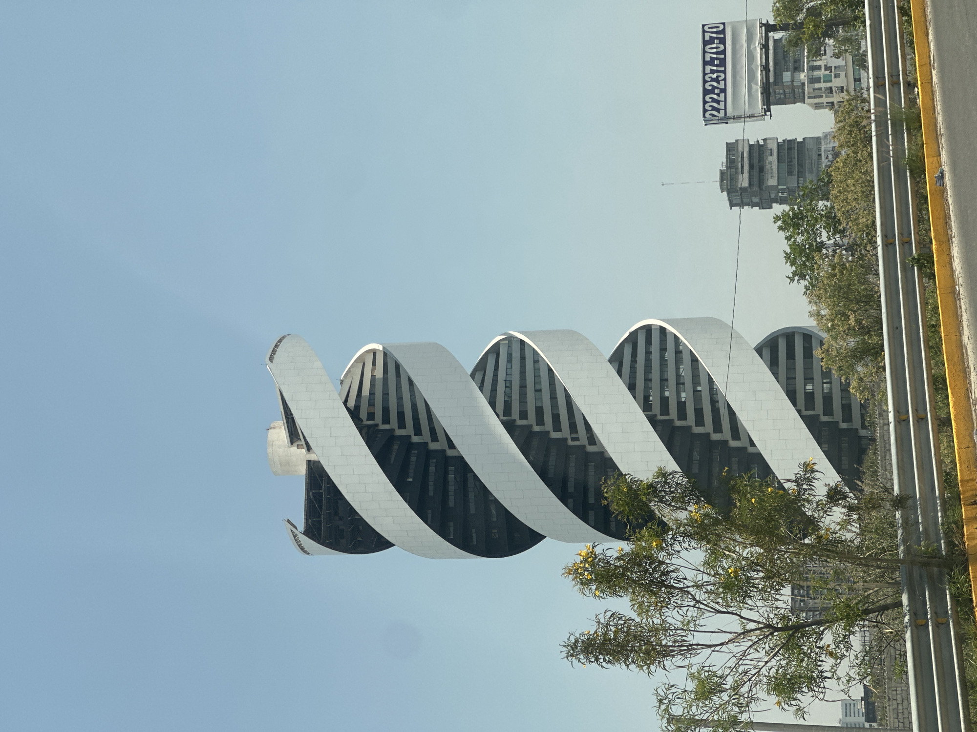 The image, oriented correctly, showcases a distinctive modern building with a unique, spiraling or ribbon-like architectural design against a clear, light blue sky in San Bernardino Tlaxcalancingo, Mexico. The building's exterior features prominent white, undulating bands that wrap around dark, reflective window sections.
In the mid-ground and background, several other buildings with more conventional high-rise designs are visible. To the right of the frame, a metal guardrail or barrier parallels a road, highlighted by a bright yellow curb or painted line. A leafy tree with small yellow flowers extends into the lower right and center of the frame, partially obscuring the main building's base. More trees and greenery are scattered throughout the scene.
The bright daylight and cloudless sky indicate a sunny day. No human activity is depicted. A billboard is visible in the distance, displaying the phone number "222-237-70-70". The scene captures an architectural landmark within its urban setting.