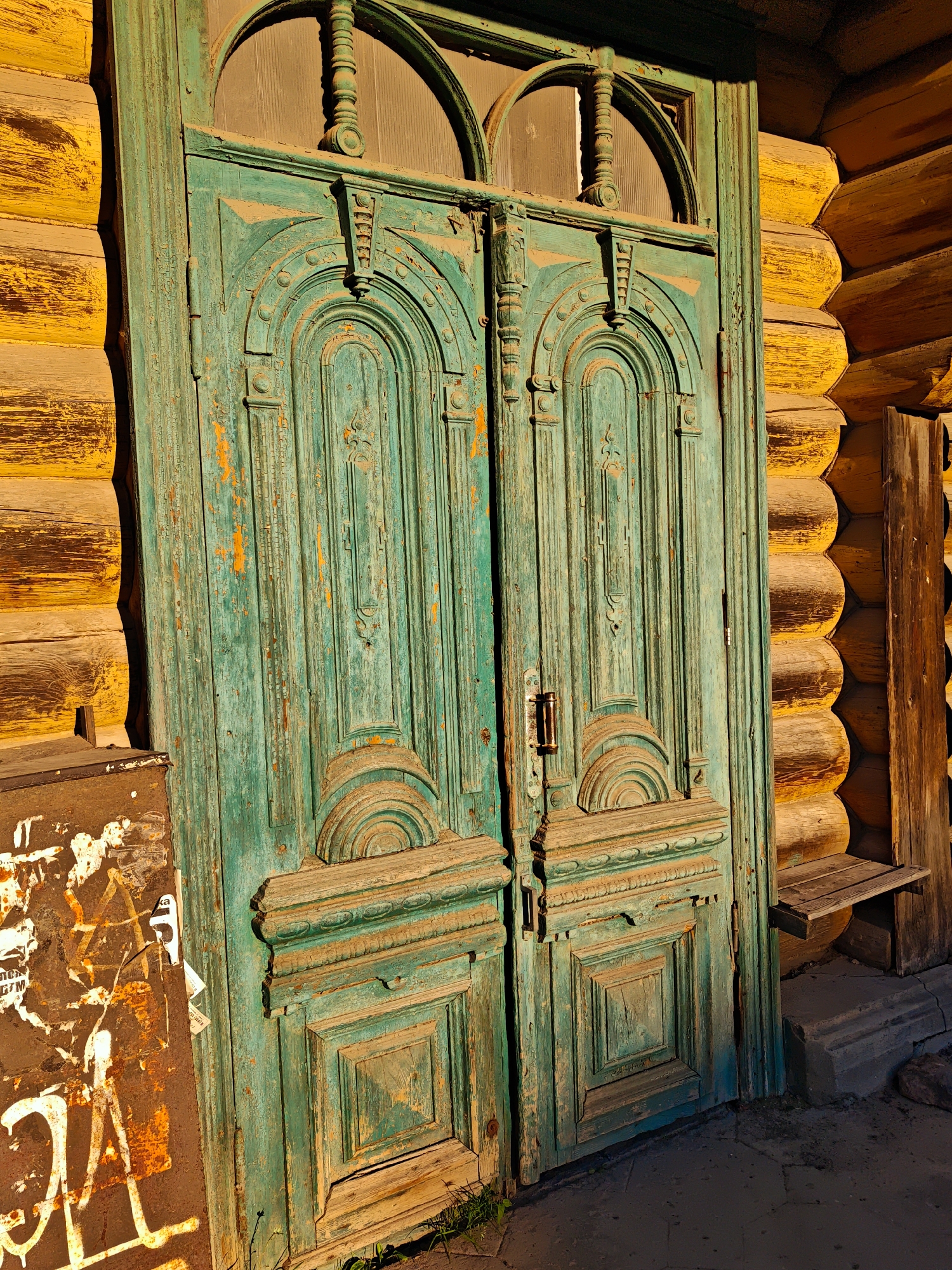 An old, turquoise-painted double door with ornate carving stands adjacent to a yellow, log-built wall. The paint on the door is chipped, revealing wood underneath, and the door itself appears weathered. The log wall has a rough texture. To the left of the door, a rusty metal surface is covered in graffiti, including white spray-painted lettering and symbols. A small wooden shelf is attached to the log wall to the right of the door. The ground in front of the door is paved with dark grey stones, and a few blades of grass are visible. The scene appears to be an exterior shot, possibly in an alley or on a street, with warm, directional lighting suggesting either morning or late afternoon sun. The location is identified as Kimry, Russia. No people are present in the image.