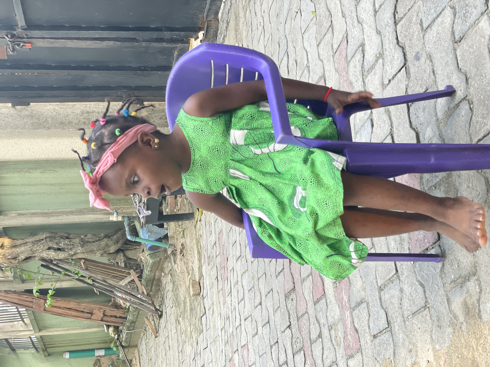 A young girl is seated outdoors in a purple plastic child's chair on interlocking grey and reddish concrete pavers in Ado, Nigeria. The girl has dark skin and wears a bright green sleeveless dress with a geometric pattern. A pink fabric headscarf or bow is tied on her head, and her dark hair is styled in small braids adorned with multicolored beads. She wears small gold heart-shaped stud earrings and a red string bracelet on her right wrist. Her bare feet rest on the paved ground. Her mouth is slightly open, and her gaze is directed upwards and to the left, indicating observation or surprise.
The background consists of a light green painted concrete wall. To the left of the girl, behind the wall, is a metal fence or grate section, a vertical green and white pipe, and a collection of wooden planks and other debris leaning against the wall. A large tree with a textured, gnarled trunk stands behind the wall in the midground. To the right of the girl, a dark metal gate secured with a red padlock is visible, alongside a light-colored concrete pillar. A small metal table, a blue cloth, and a segment of a blue pipe are visible near the ground on the right side of the frame.