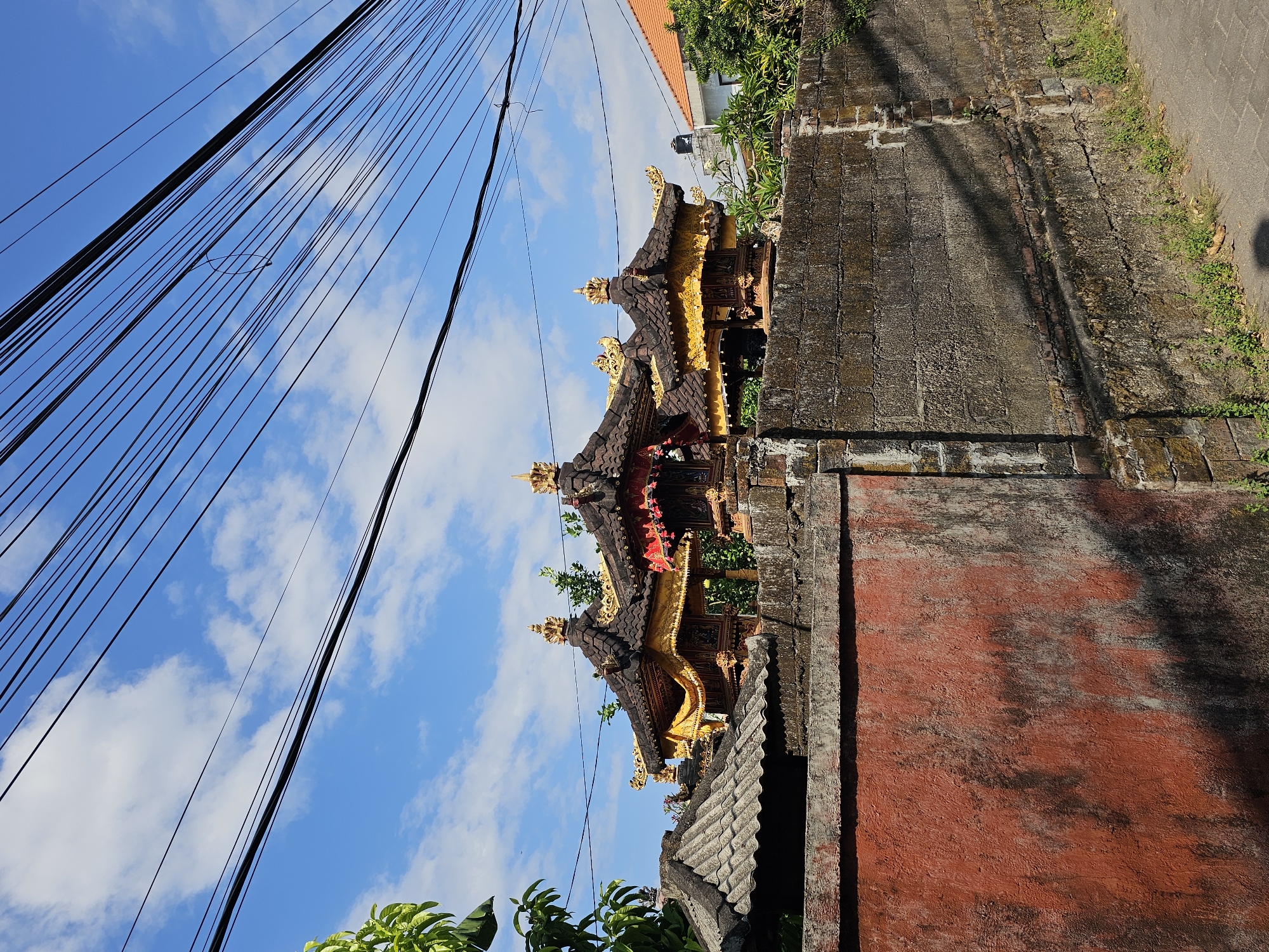 This is a medium-shot, eye-level, outdoor photograph taken in Denpasar, Indonesia, featuring a traditional Balinese structure. The scene is dominated by the sky, filled with a blue expanse and scattered white clouds, with a prominent network of overhead electrical wires crisscrossing the upper portion of the frame.
In the mid-ground, behind a weathered stone and concrete wall, sits a tiered, ornate structure. The structure has multiple dark, tiled roofs adorned with golden decorations and embellishments. Yellow and red fabric drapes are visible, adding to the decorative appeal. The building appears to be a shrine or temple, characteristic of Balinese architecture.
The wall in the foreground is a mix of textures and colors. The left section is a rough, red-painted surface, showing signs of wear and tear with dark patches and a textured finish. To the right, the wall transitions to a rougher, gray stone and concrete construction, with visible bricks in places and patches of moss and small plants growing along its base.
To the left of the frame, the lush green leaves of a tree extend into the shot from the upper left corner. In the background, glimpses of residential buildings with orange tiled roofs and green vegetation can be seen, suggesting a semi-urban or residential area. The ground in the foreground consists of a paved sidewalk or pathway. No people are visible in the image. The lighting suggests it is daytime, with shadows cast on the wall, indicating sunlight.
