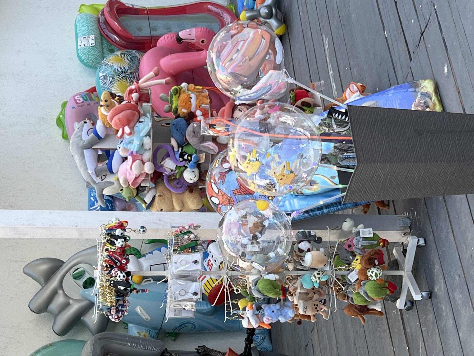 This image depicts an outdoor market stall in Năvodari, Romania, likely near the Black Sea coast, given the presence of inflatable beach toys. The stall displays a variety of merchandise, including plush toys, keychains, and novelty balloons. In the foreground, a white multi-tiered rack is filled with numerous small plush animal keychains, some resembling characters like bubble tea. Above these, a display rack holds various keychains with gold rings and colorful charms, including what appear to be sports-themed designs. Clear, large balloons filled with confetti and featuring cartoon characters like Spider-Man and Pikachu are prominently displayed. In the background, a collection of large inflatable items, including pool floats and possibly a beach ball, are visible, some with vibrant patterns. The stall is set up on a wooden deck or boardwalk, suggesting a casual, recreational setting. No people are visible in the frame.