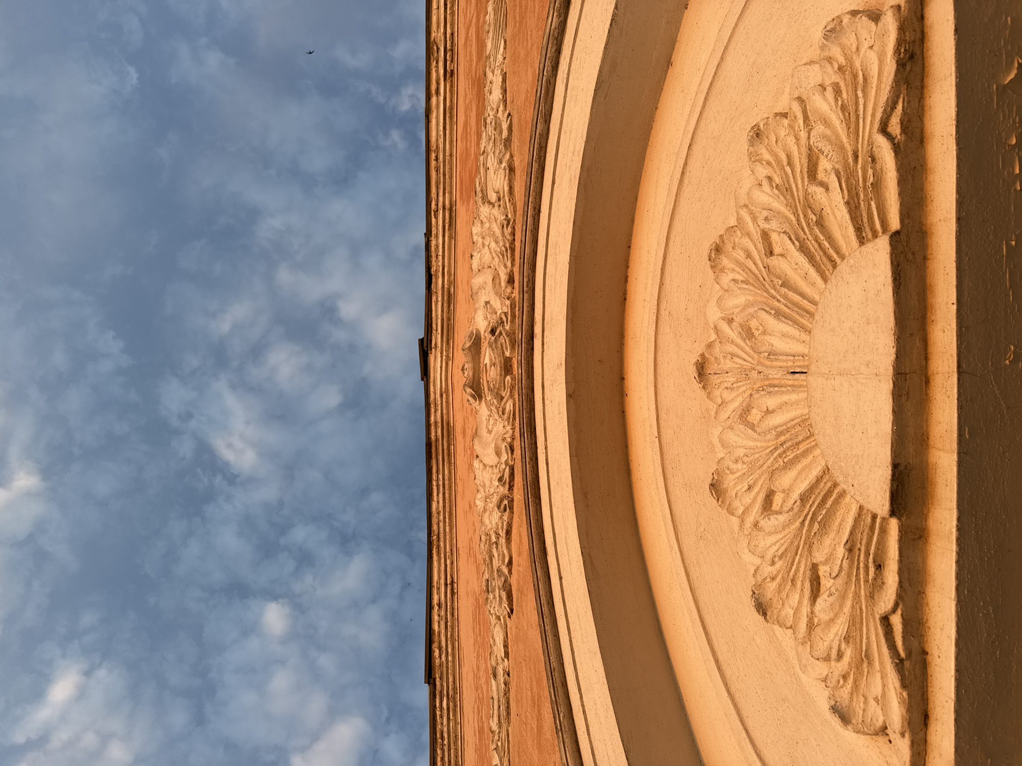 The image shows a section of an ornate, likely historical, building against a sky with scattered clouds. The building features detailed relief work with a floral-like pattern in the center, framed by an arch. The building appears to be made of a light-colored material, possibly stone or stucco, with a warm, possibly sunset-related, lighting. A bird is visible in the sky. It is unknown if the building is in the city of Votkinsk, Russia.