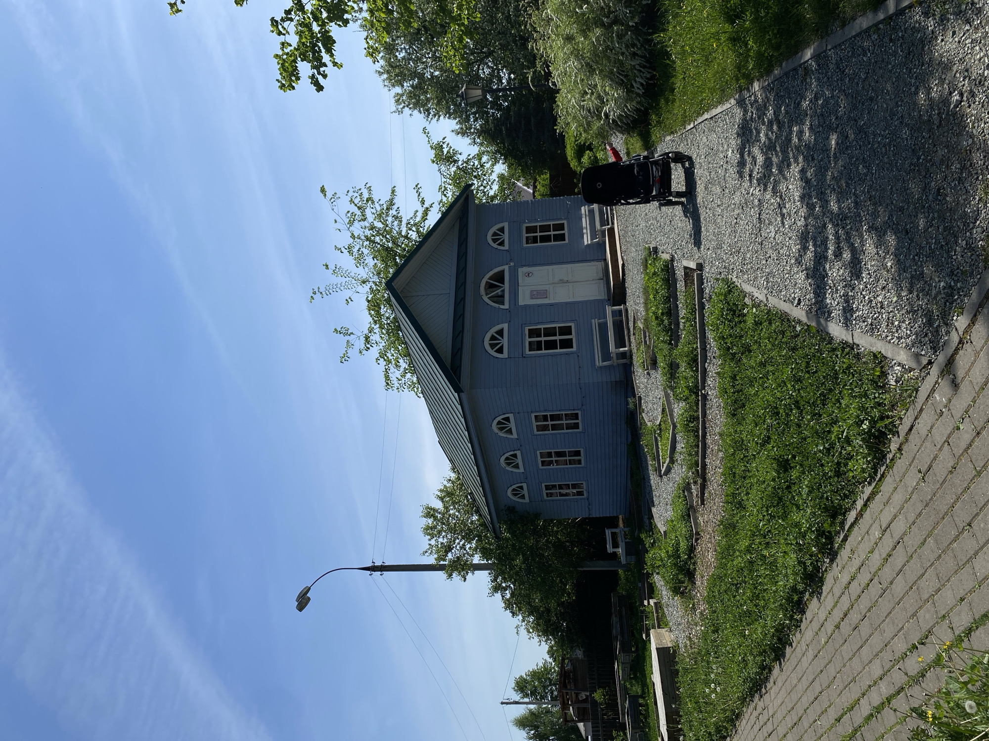 The image shows a light blue, two-story wooden house with white trim and a green roof located in Votkinsk, Russia. The house has a symmetrical facade with a central door, arched windows on the upper floor, and rectangular windows on the lower floor. A small tree appears to be growing directly in front of the roof. In front of the house is a gravel pathway leading from a stone brick pathway near the edge of the road. To the right of the house along the walkway is a black stroller. A street lamp is visible to the left of the house. The sky is blue with light cirrus clouds. The surrounding environment includes grass, greenery, and trees, indicative of a residential area. Utility poles are visible in the background.