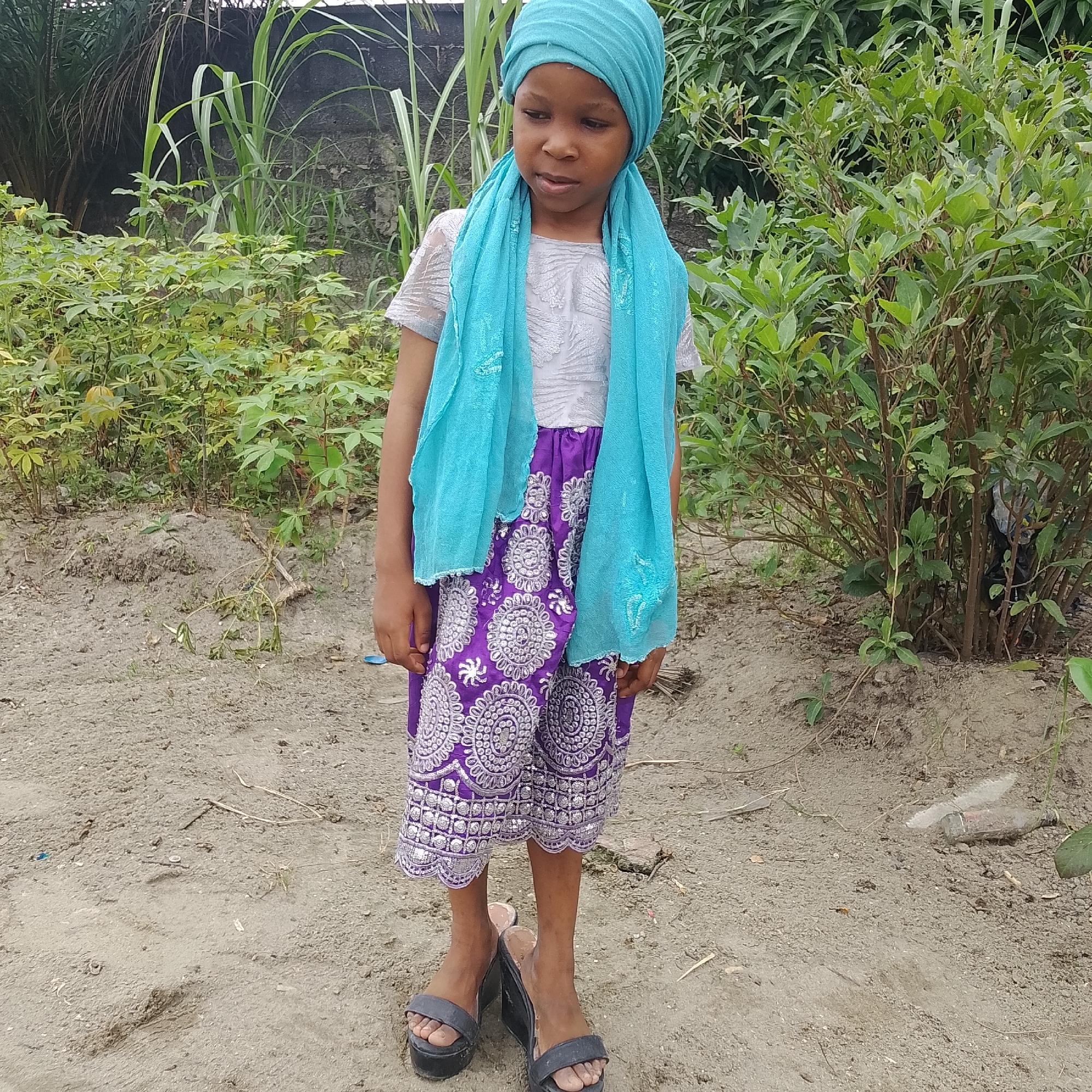 In Ado, Nigeria, a young girl stands in a sandy, outdoor area with dense green foliage behind her. She is wearing a light grey short-sleeved shirt with a white, textured overlay, a vibrant purple skirt with intricate silver circular patterns, and a bright turquoise headscarf and matching scarf draped around her neck and shoulders. Her feet are clad in dark-colored platform wedge sandals. The ground is composed of light brown sand with scattered dry leaves and small twigs. In the background, a rough, dark wall is visible behind tall, green stalks of sugarcane. The overall impression is of a child in a natural, possibly rural, setting within the city.