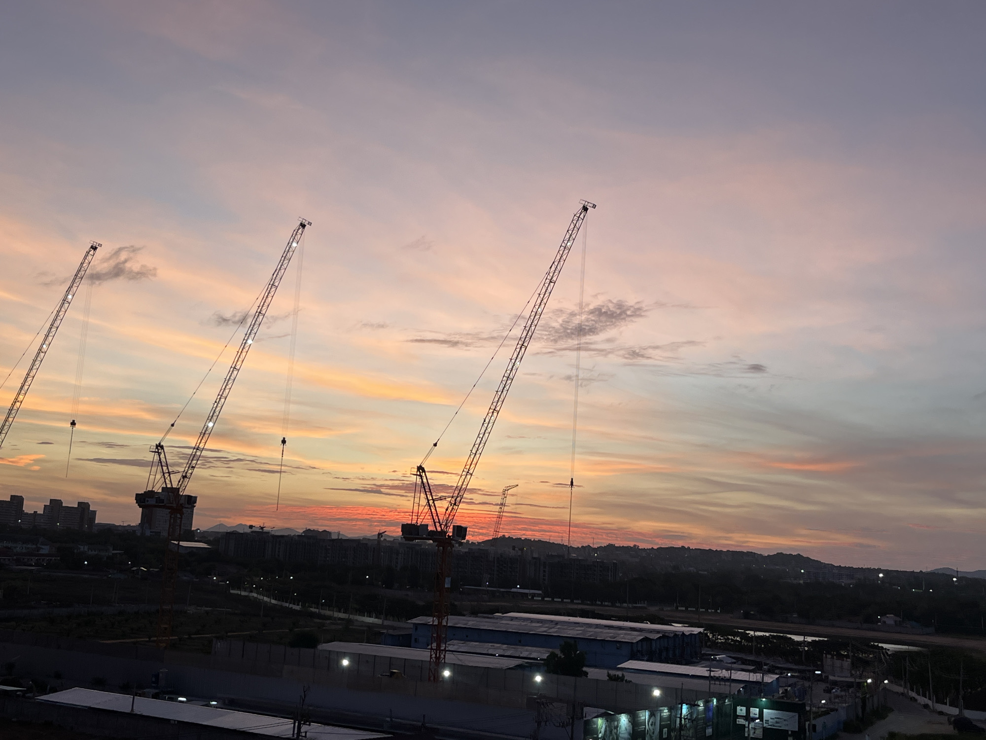 The image shows a cityscape at dusk in Pattaya City, Thailand. The sky is a mix of pink, orange, and light purple hues. Several construction cranes are silhouetted against the colorful sky. In the background, there are buildings and a hilly landscape. In the foreground, there are low-rise buildings and structures with lights on. There are no people visible in the image.