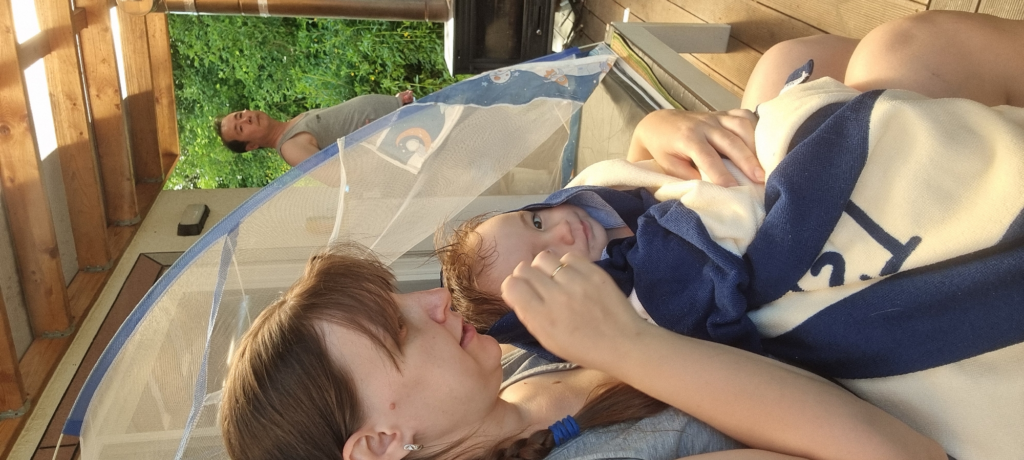 In Dunnae-myeon, South Korea, a woman with brown hair, partially braided with a blue hair tie, lies in what appears to be an outdoor lounging area. She is covered with a blue and white striped towel and is holding a child wrapped in the same towel. Above them, a man lies within the confines of a blue-trimmed mosquito net. The area is partially shaded by a wooden structure. Outside the structure, green foliage is visible.