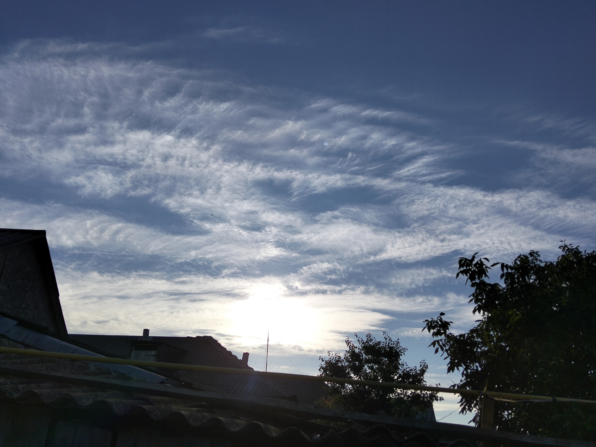 The image shows a silhouetted view of rooftops and trees against a sky filled with cirrus clouds. The sun is bright and positioned in the center of the frame, partially obscured by clouds. The sky is a deep blue, transitioning to lighter shades near the horizon. The foreground is dominated by dark, textured rooftops, with a yellow pipe running horizontally across the scene. A tall, thin antenna-like structure is visible behind the rooftops, and to the right, the dark silhouettes of trees are prominent against the bright sky. The overall scene suggests an outdoor environment, possibly a residential area at dawn or dusk.