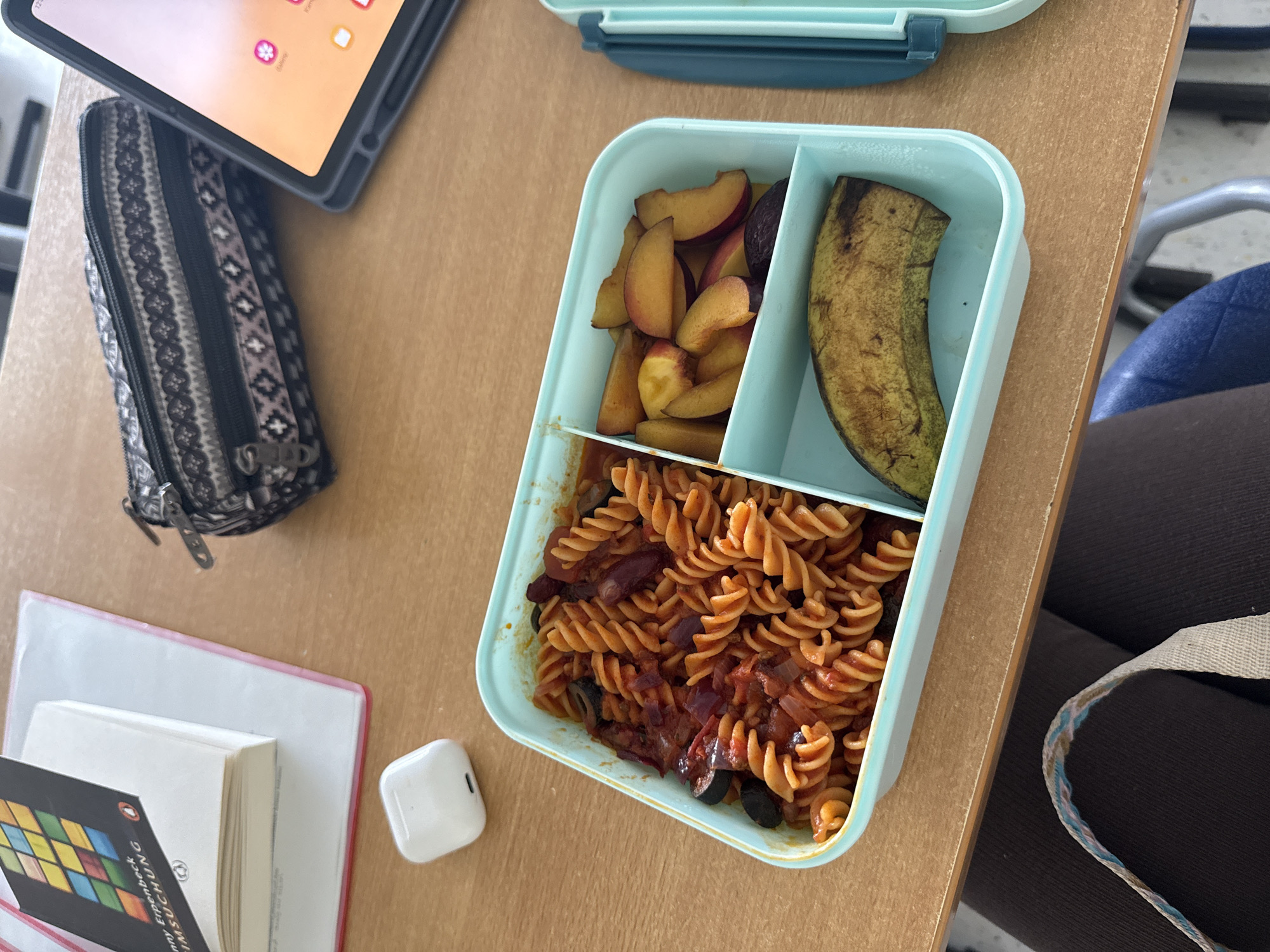 The image shows a close-up view of a wooden desk with various items. In the foreground, a light blue lunchbox is divided into three compartments. The largest compartment contains rotini pasta with a red sauce, olives, and possibly beans or red onion. The second compartment holds sliced peaches. The third compartment contains a whole, ripe banana. To the left of the lunchbox, a white wireless earbud case is visible. Further left, a book with a colorful geometric cover and text in German is partially open. Behind the lunchbox, a patterned pencil case and a tablet device are placed on the desk. The desk surface appears to be made of wood. The lighting suggests an indoor setting. There is no explicit indication of the location being the city of Hanau, Germany, beyond the prompt's directive, and no people are visible.