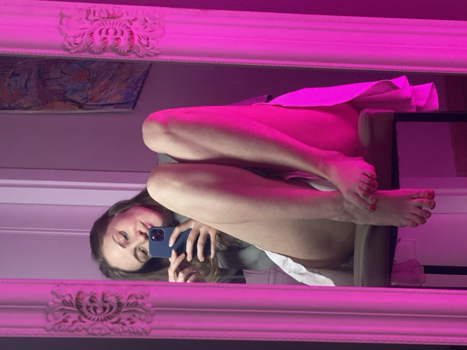 A woman is reflected in a mirror, holding a phone to take a picture. She sits on a chair, legs crossed, with a white cloth draped over them. Her face is visible as she looks into the phone's camera. The mirror's ornate frame is illuminated with a pink light, which casts a magenta hue over the scene. In the background, a glimpse of artwork hangs on the wall.