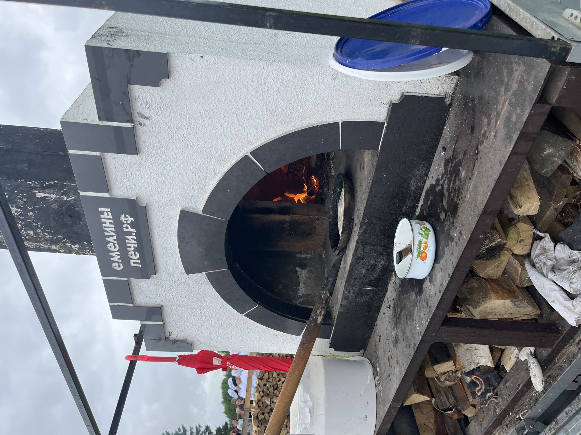 An outdoor cooking scene features a white and black brick oven, stylized like a castle turret, positioned on a dark workbench. The oven has an arched opening, inside which a wood fire burns brightly on the left, illuminating a dark frying pan containing a light-colored disc, possibly cooking food. A long wooden paddle with a dark, possibly metal, end is extended into the oven, resting on the pan. Above the oven's arch, the Russian words "ЕМЕЛИНЫ" and "ПЕЧИ.РФ" are displayed in white lettering on black plaques.
To the left of the oven, a large white plastic bucket containing a white liquid or batter is visible, with the wooden paddle handle submerged. Below the workbench, a pile of split firewood is neatly stacked. On the right side of the workbench, a small white enamel pot with a colorful fruit and leaf pattern holds a dark utensil. Adjacent to the oven's right side, a blue plastic lid and a white plastic lid lean against a metal support structure.
In the mid-ground on the left, behind a wooden barrier, a group of people, some wearing white chef hats, are visible. The sky overhead is overcast and gray. A red umbrella is tied to an overhead black metal bar, which forms part of a frame supporting the setup. The overall scene suggests a temporary outdoor cooking demonstration or event.