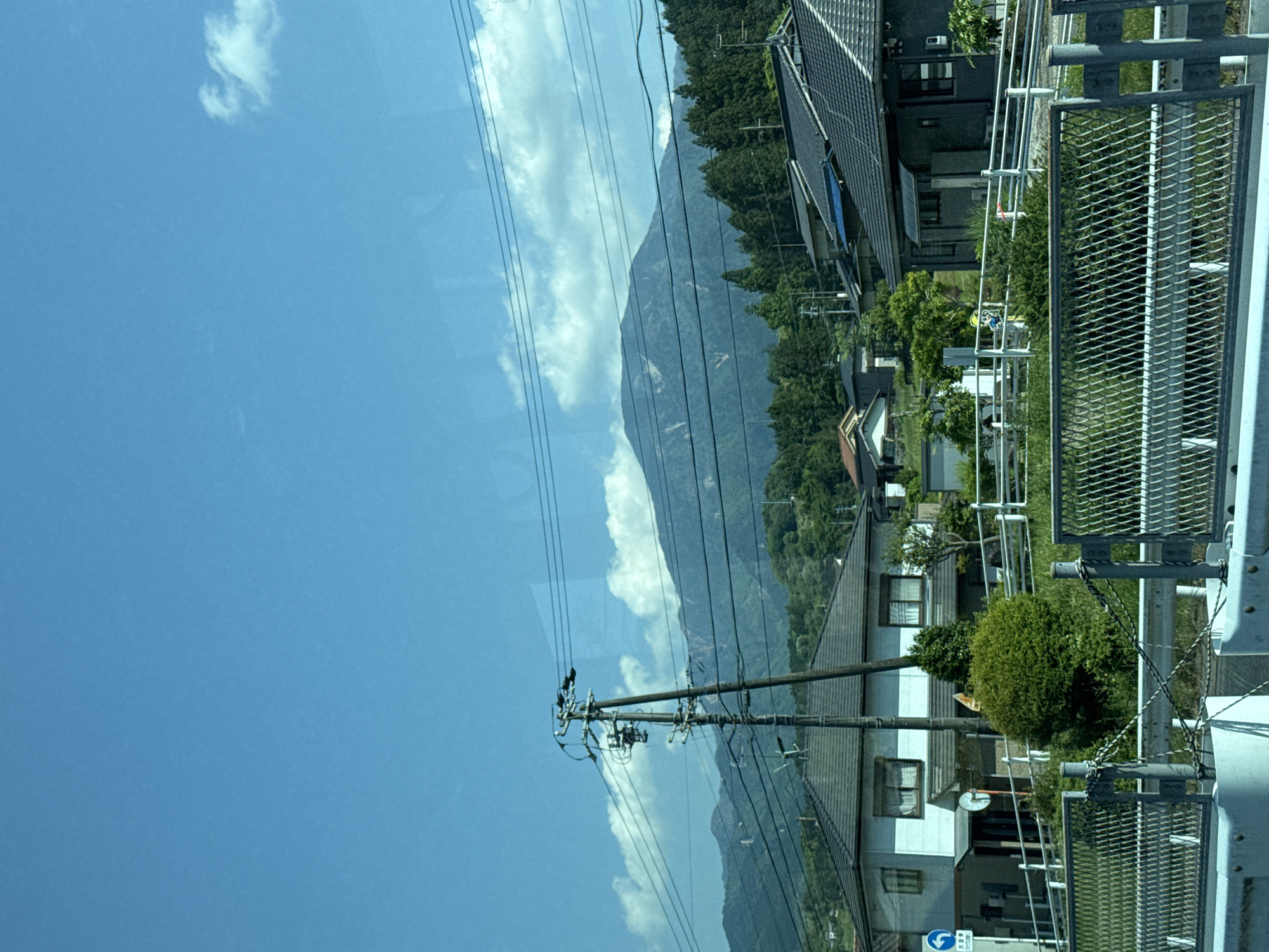 The image depicts a roadside view in Nakatsugawa, Japan. A mountain range is visible in the background, partially obscured by clouds. In the foreground, power lines stretch across the sky, running parallel to the road. Houses and trees line the road. A metal guardrail runs along the side of the road.