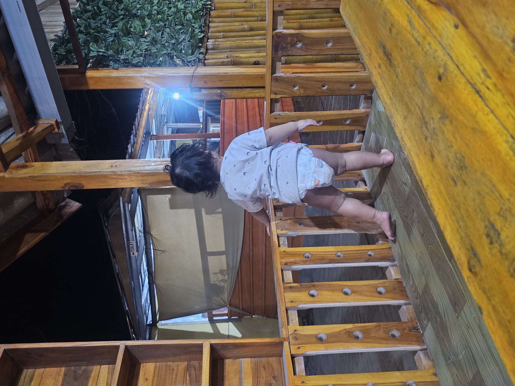 The image captures a child, from behind, standing near a wooden railing on a paved floor. The child is wearing a light-colored t-shirt and a diaper. The environment appears to be an outdoor seating area, possibly a restaurant or cafe, constructed with wood and bamboo elements. A bright light source is visible in the background, and lush green foliage can be seen on the right side of the frame. A sign that reads "TOILET" is also visible in the distance. The scene is likely situated in Jenu, Indonesia, given the context provided. The overall impression is a casual, nighttime setting.
