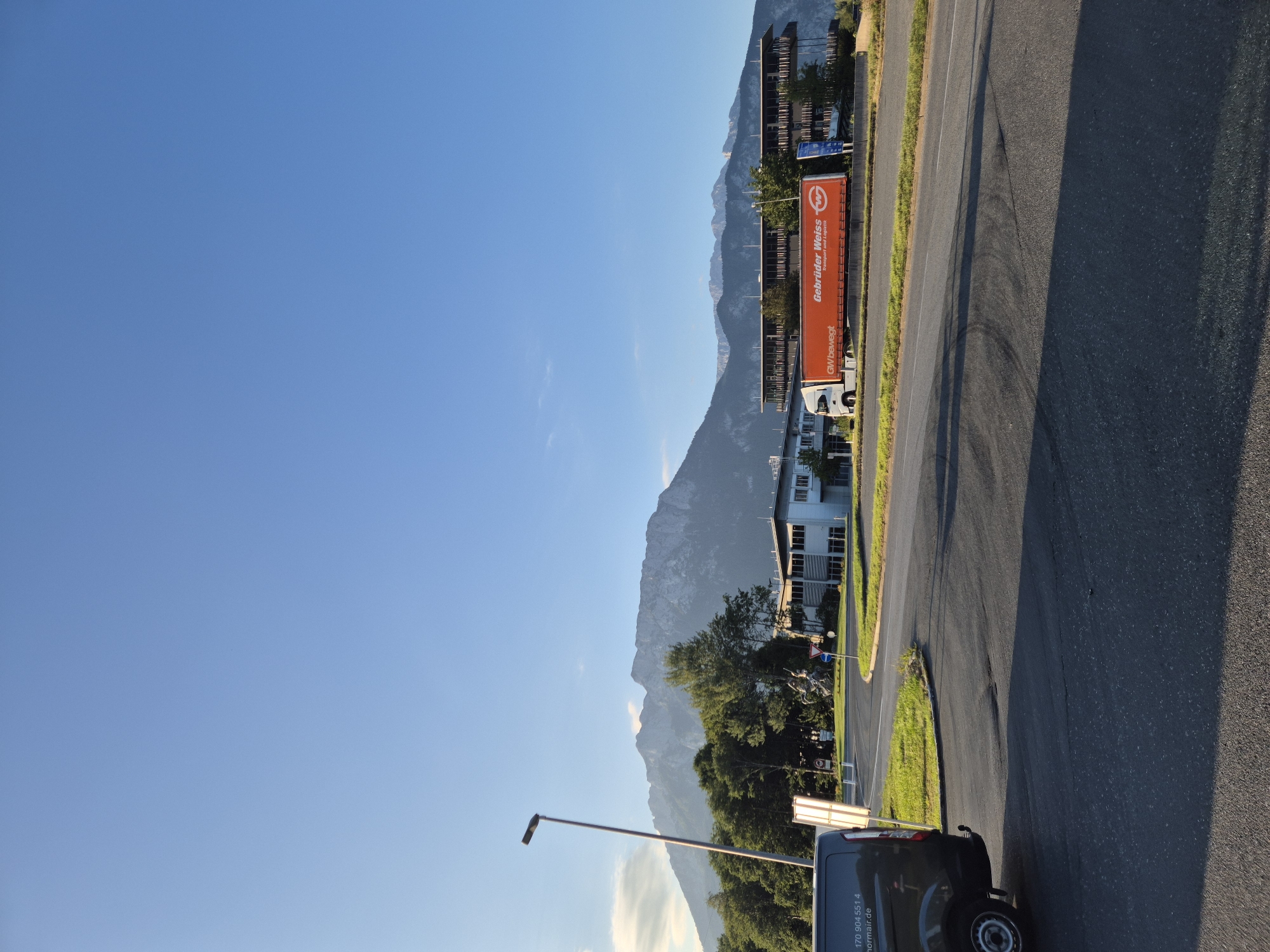 The image captures a scene in the city of Niederndorf, Austria, under a clear blue sky. In the foreground, a dark-colored van is parked on the left side of the frame. To the right, a paved road curves into the distance, with a grassy median strip separating it from another road.
Dominating the middle ground are several buildings. On the left, a light-colored building with numerous windows is visible. Further to the right, a larger building with a distinctive cantilevered upper level, featuring a facade of vertical fins, stands out. Between these structures, a white semi-trailer truck with an orange cargo box is parked. The truck displays the text "Gebrüder Weiss Transport und Logistik" and the "GW" logo. Adjacent to the truck, trees and greenery are present. A statue of a figure on horseback is also visible near the buildings.
In the background, a majestic mountain range stretches across the horizon, its peaks partially illuminated by sunlight. A single street lamp stands tall on the left side of the image, near the van. The overall impression is one of an industrial or commercial area situated in a scenic location, with the imposing mountains providing a dramatic backdrop.