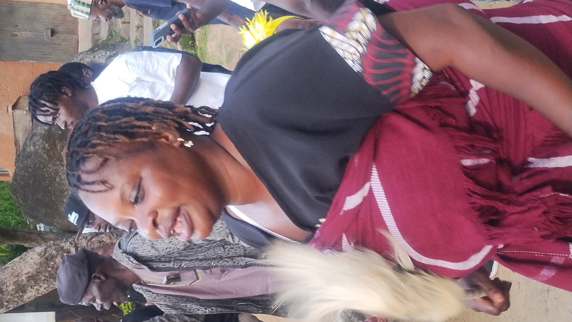 A medium shot shows a woman in the foreground, facing left and looking upwards, her mouth slightly open as if speaking or singing. She has dark skin, dark dreadlocks, and is wearing gold earrings. She is draped in a dark top over a maroon and white patterned cloth with fringes. A plume of white feathers, possibly part of a ceremonial outfit, is held in her left hand, extending upwards.
Behind her, to the left, an older man with dark skin and grey hair, wearing a dark cap and a patterned jacket over a light pink garment, looks towards the left. Another man, wearing a black baseball cap, is visible between the older man and the woman.
To the right of the woman, a man with dark skin and dreadlocks, wearing a white t-shirt, looks to his left. Further back and to the right, another man wearing a colorful patterned hat, with grey facial hair, is visible. A dark-skinned man holding a mobile phone is also present to the right.
The background suggests an outdoor setting with a rough, textured wall or structure on the right, possibly part of a building, with a wooden door and steps leading upwards. A large rock formation is visible behind the individuals, and foliage is present in the upper left. The ground appears to be dirt or earth.
The scene appears to be a public gathering, potentially a cultural event or celebration. The specific location is stated to be in Jos, Nigeria.