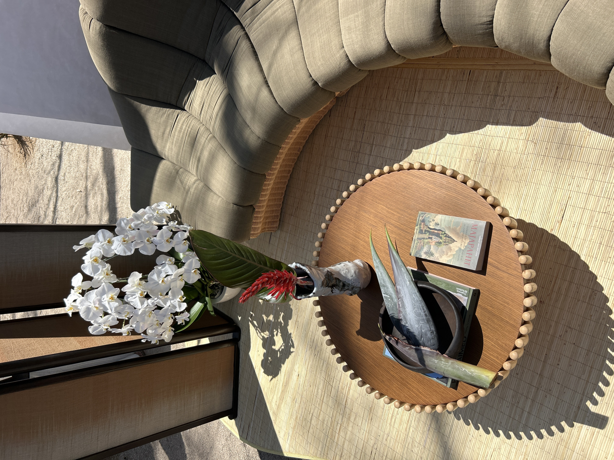 A round wooden coffee table with beaded edges sits on a woven mat. On the table are two books, a dark bowl with two large green leaves, and a vase holding a red ginger flower and a green leaf. To the right of the table is a large, curved sofa with olive-green upholstered cushions and a rattan base. Behind the table, to the left, is a tall, wooden folding screen with fabric panels. White orchids in a white pot are visible behind the folding screen. The scene appears to be an outdoor setting, with shadows cast by sunlight from above. The location is described as Denpasar, Indonesia.
