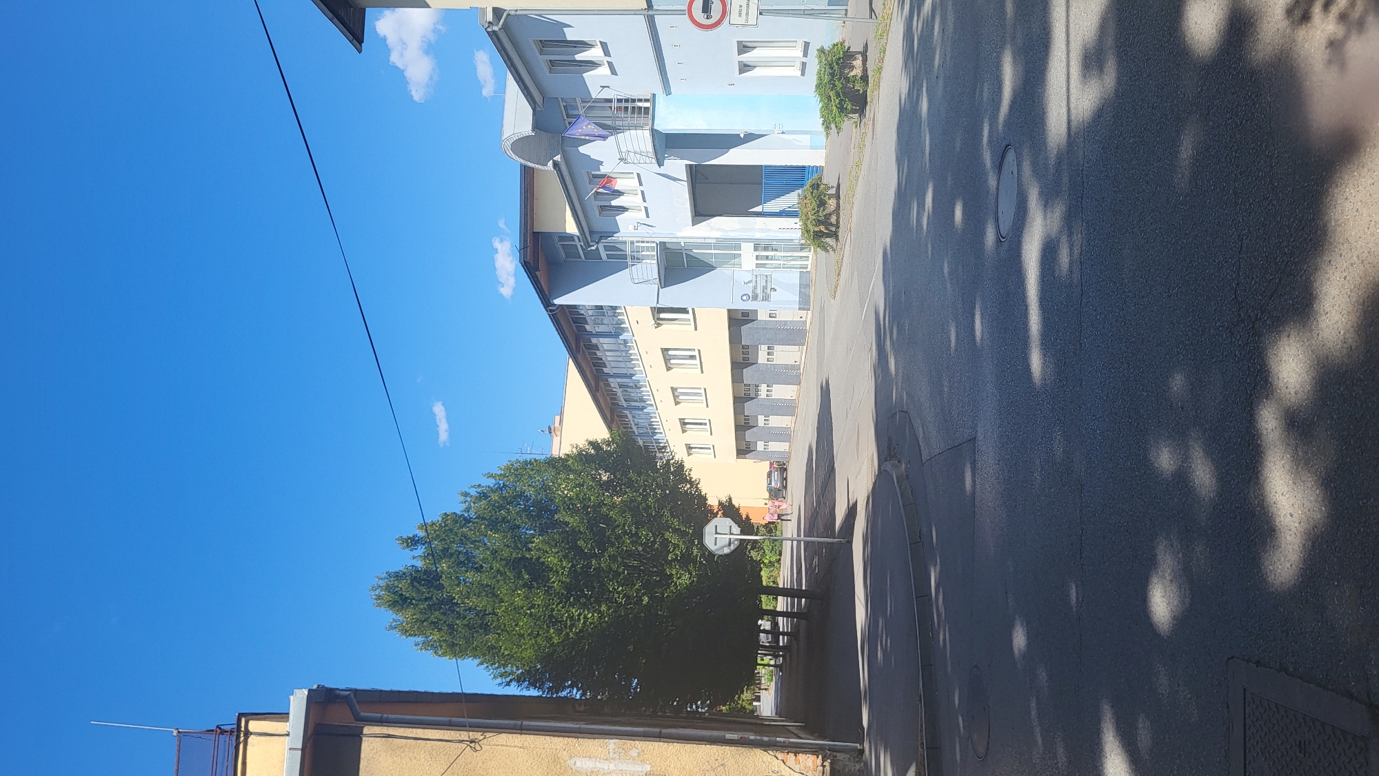 The image depicts a street scene in Prešov, Slovakia, on a bright, sunny day. In the foreground, a paved road is visible with shadows of trees cast across it, suggesting it is midday. On the right side of the road, a light blue building stands with multiple garage doors on the ground floor and windows on the upper levels. Two flags, one Slovak and one European Union flag, are mounted on a balcony on the blue building. Adjacent to the blue building is a yellow building with a similar architectural style. On the left side of the street, several tall, mature trees line a sidewalk. A yellow building with rough textured walls is partially visible on the far left. A traffic sign with a red circle and a horizontal bar, indicating a prohibition, is visible near the blue building. Further down the street, a few people are walking, and a car is parked. The sky is clear and blue with a few small white clouds.
