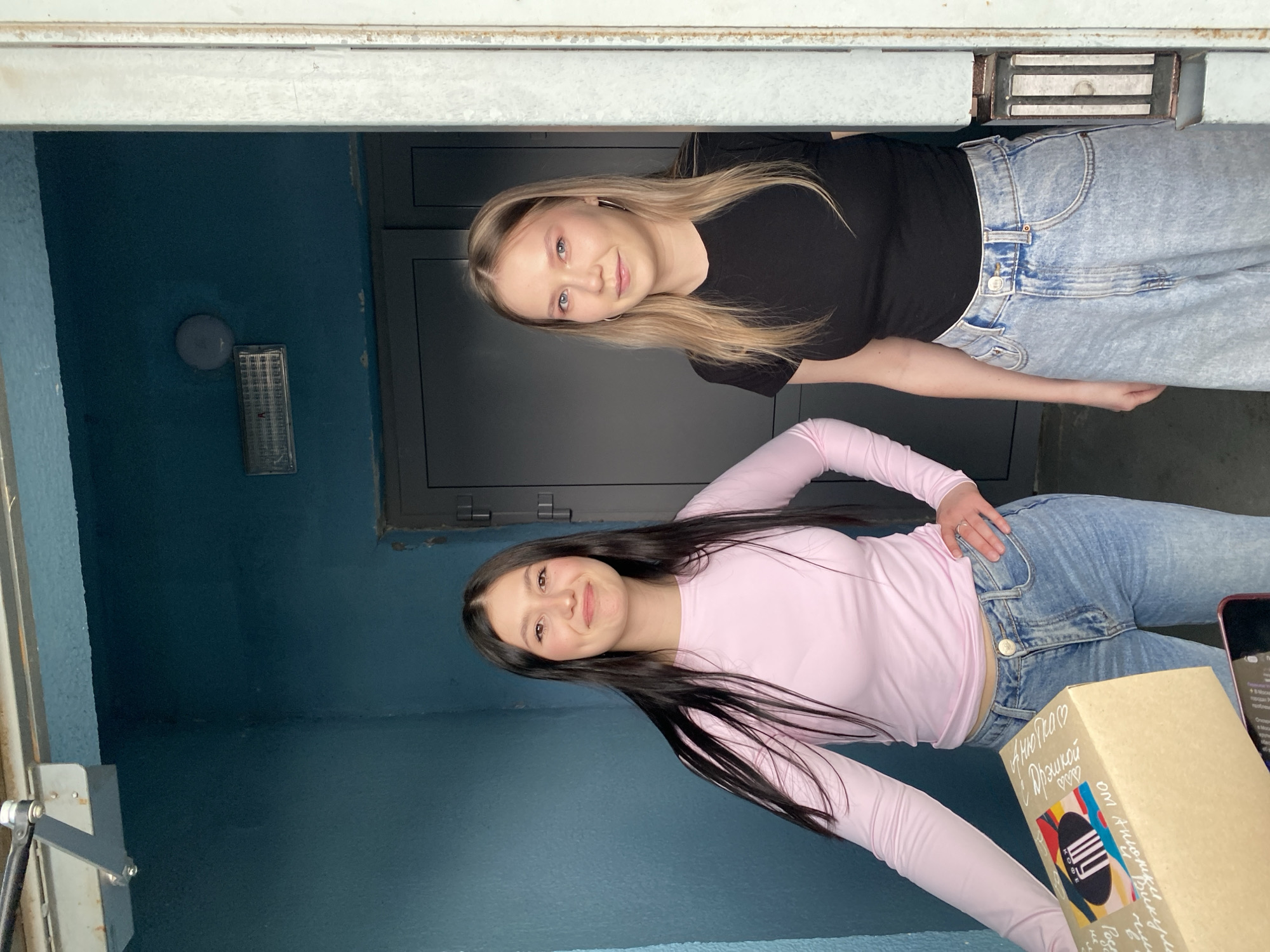 Two young adult females are depicted standing in an indoor entrance or hallway in Chaikovsky, Russia. The individual on the left, with long dark hair, wears a light pink long-sleeved top and light blue jeans. She smiles towards the camera while holding a light brown cardboard package. The package is decorated with colorful abstract designs and Cyrillic text, including "КОФЕ" (Coffee), "С днем рождения!" (Happy Birthday!), and "от Анюты и Викули" (from Anya and Vika). A red-cased smartphone is partially visible beneath the package. The individual on the right, with long blonde hair and silver hoop earrings, wears a black short-sleeved top and light blue wide-leg jeans, and looks towards the camera with a neutral expression. The background consists of textured dark teal walls. A dark gray door is situated behind the right individual. To the far right, a light-colored door frame with a vertical metallic magnetic lock plate is visible. Overhead, a silver door closer mechanism, a circular wall light fixture, and a rectangular fluorescent light fixture are present.