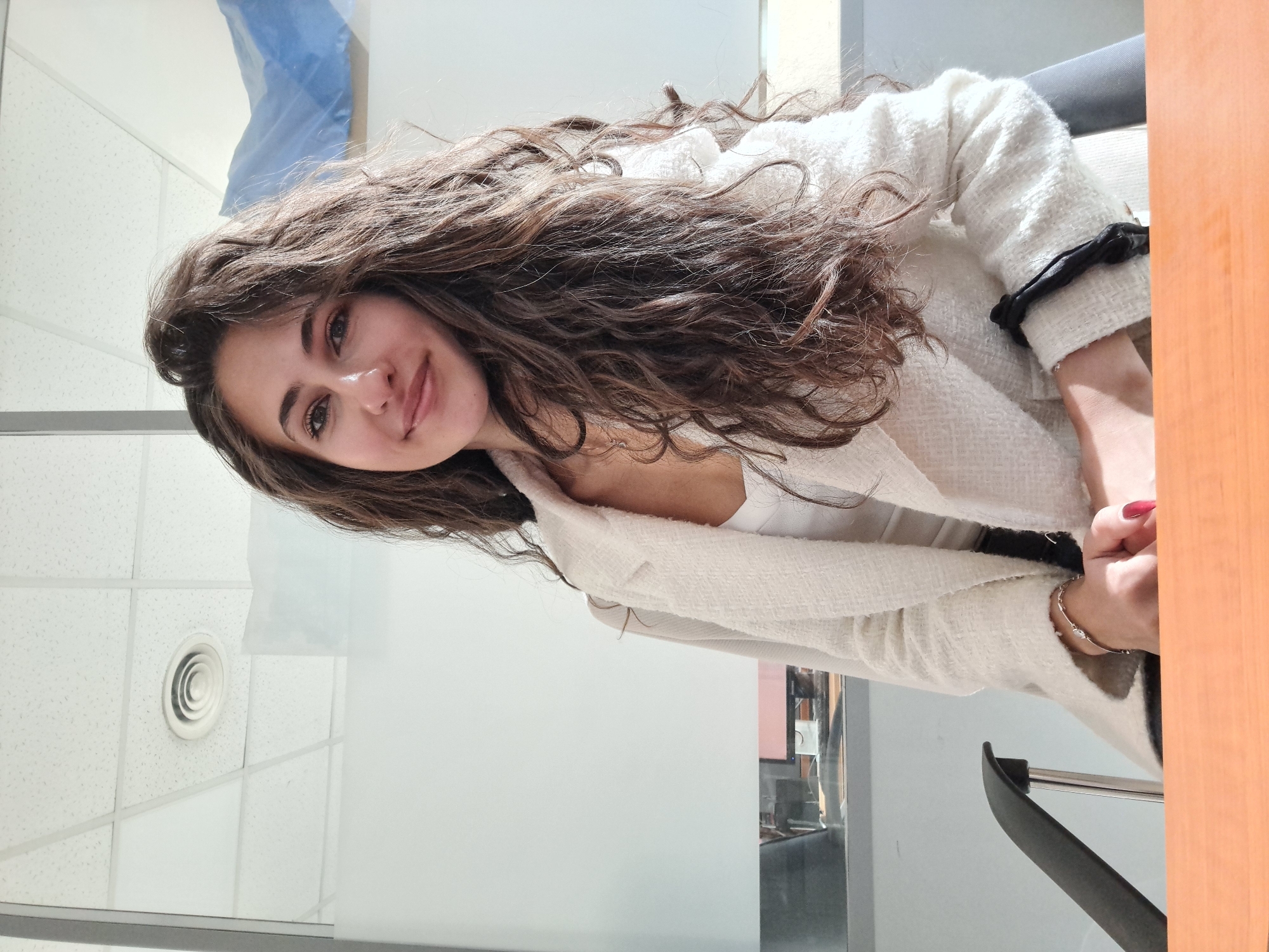 A young woman with long, wavy brown hair is posing for a portrait indoors. She is wearing a white tweed jacket over a white top. She has a bracelet on her right wrist and red nail polish. The background appears to be an office space with a white tiled ceiling with circular air vents, white walls, and glass partitions. The image does not contain enough information to determine the exact location in Sofia, Bulgaria.