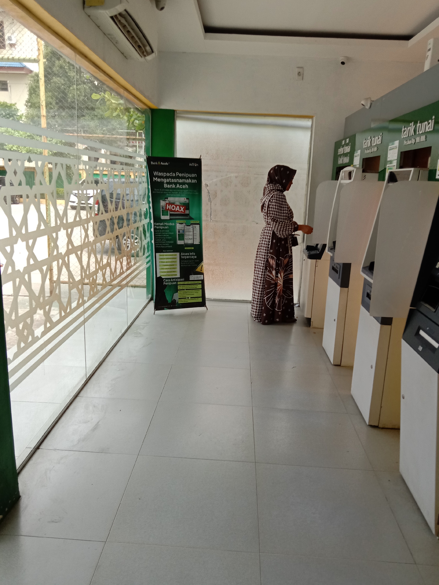 A woman is at a bank ATM in Lhokseumawe, Indonesia. The woman is wearing a patterned brown and black robe and a brown hijab. The bank has several ATMs, which are white. There is a sign warning of fraud in the name of Bank Aceh. The floor is tiled, and there is a large glass window to the left of the ATMs. There is a security camera on the ceiling. The bank is well-lit. Outside, there are buildings and trees.