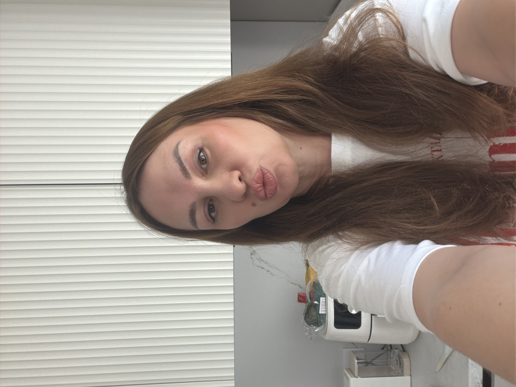 The image shows a selfie of a woman with long brown hair, taken in what appears to be a kitchen. She is wearing a white t-shirt and has her lips puckered. In the background, there are white cabinets with vertical blinds, a marble countertop, and a few kitchen appliances, including a white air fryer. The woman's face is the primary focus of the image, with her expression suggesting a playful or flirtatious mood. The lighting is bright, casting a slight shadow on the right side of her face. There is no specific indication of the city of Malinovka, Russia, or any other identifiable urban location.