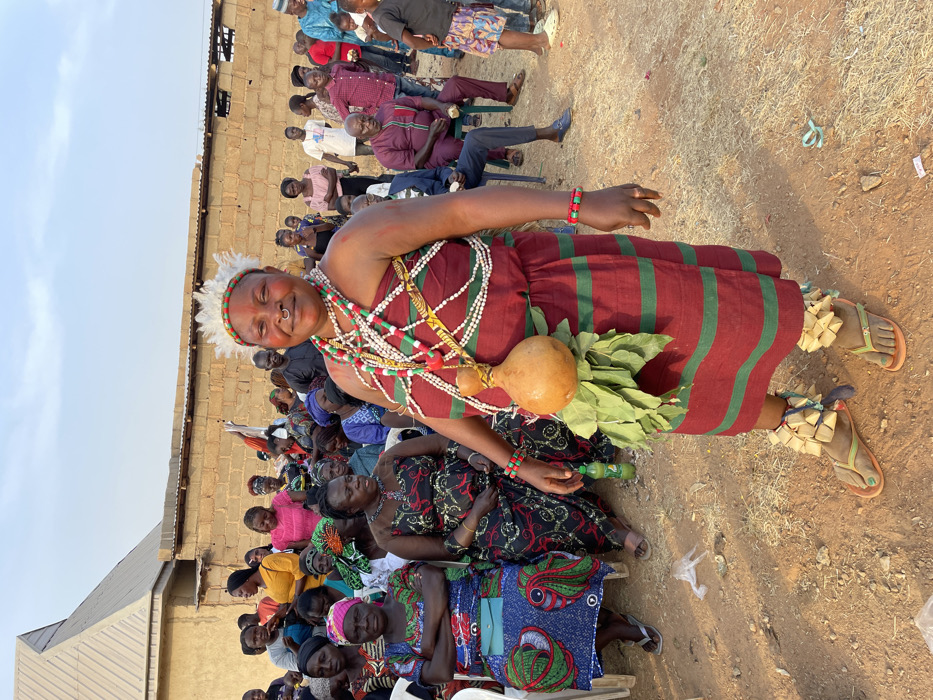 A woman stands in traditional attire, featuring a red and green striped dress, numerous colorful beaded necklaces, a gourd and greenery attached to her waist, and unique ankle accessories. A ring adorns her nose, and she wears a feathered headdress. She is surrounded by a crowd of people seated in chairs, and the setting appears to be outdoors in a rural area, with a building visible in the background.