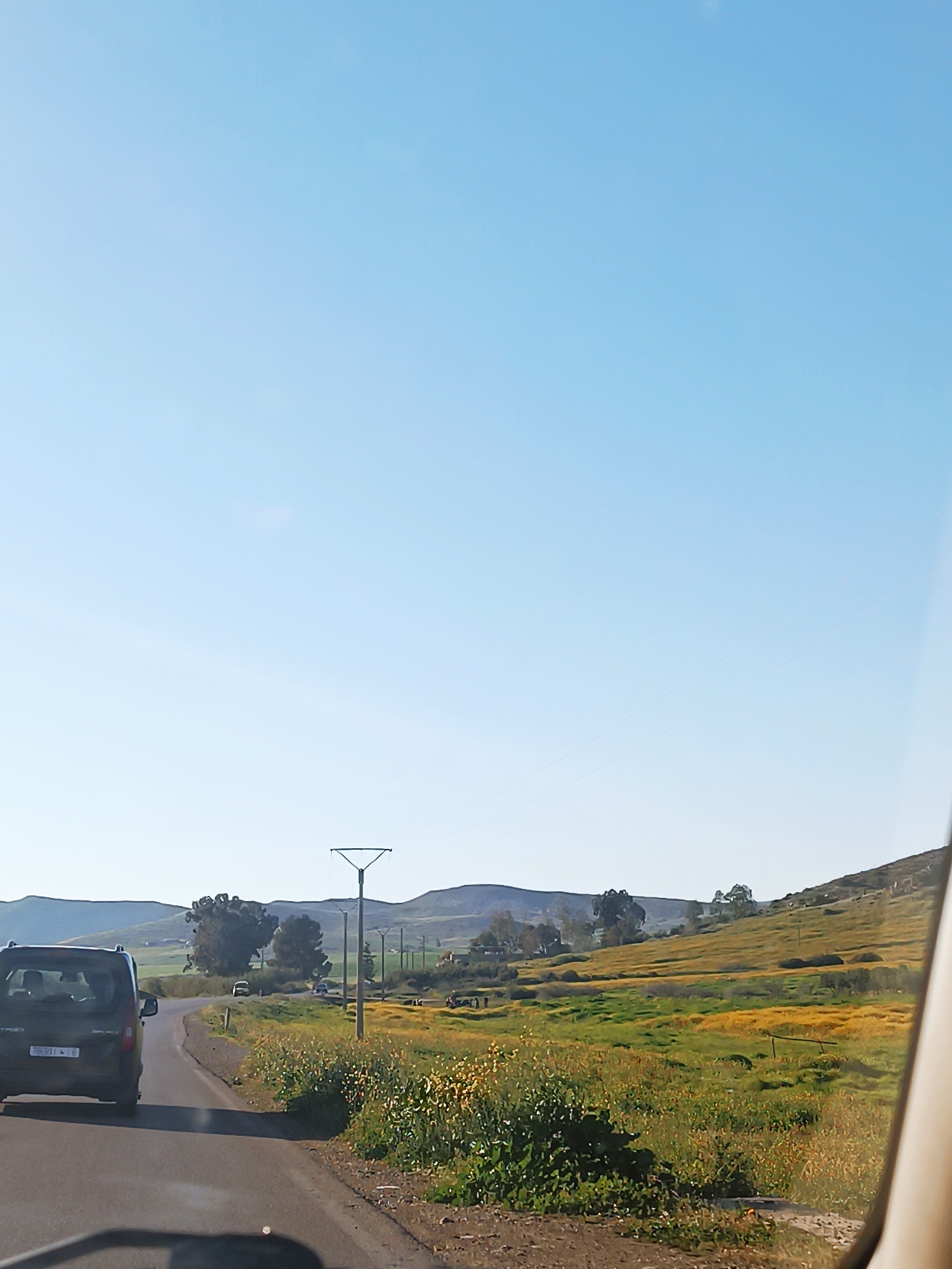 The image depicts a road scene in El Hammam, Morocco. A dark-colored van is driving on the left side of the road. Beyond the roadside, a field of green and yellow vegetation stretches towards rolling hills in the background. Overhead power lines are visible. The sky is clear and blue.