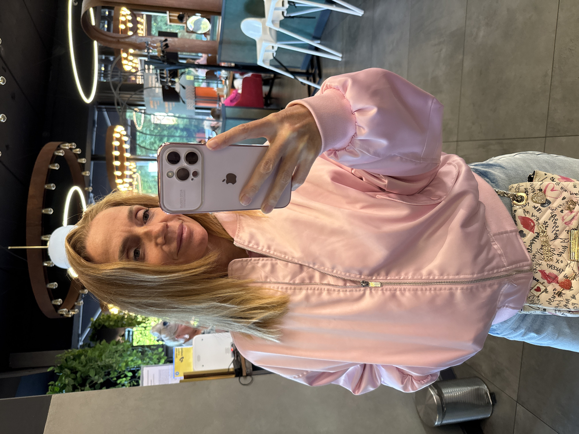 A woman with blonde hair is taking a selfie in what appears to be a cafe or restaurant. She is wearing a light pink bomber jacket and light blue jeans, and a small patterned purse is visible hanging from her belt loop. In the background, there are other patrons, tables, chairs, and decorative lighting fixtures. The environment suggests an indoor public space. There is no indication of this location being in Veliky Novgorod, Russia.