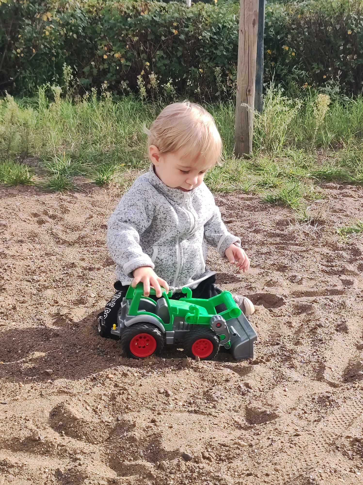 An infant with blond hair is kneeling in the sand, playing with a green and gray toy tractor. The child is wearing a gray jacket and black pants. The background consists of grass and bushes. The image appears to be taken outdoors during the daytime. The location is not specified as being in the city of Chaikovsky, Russia.