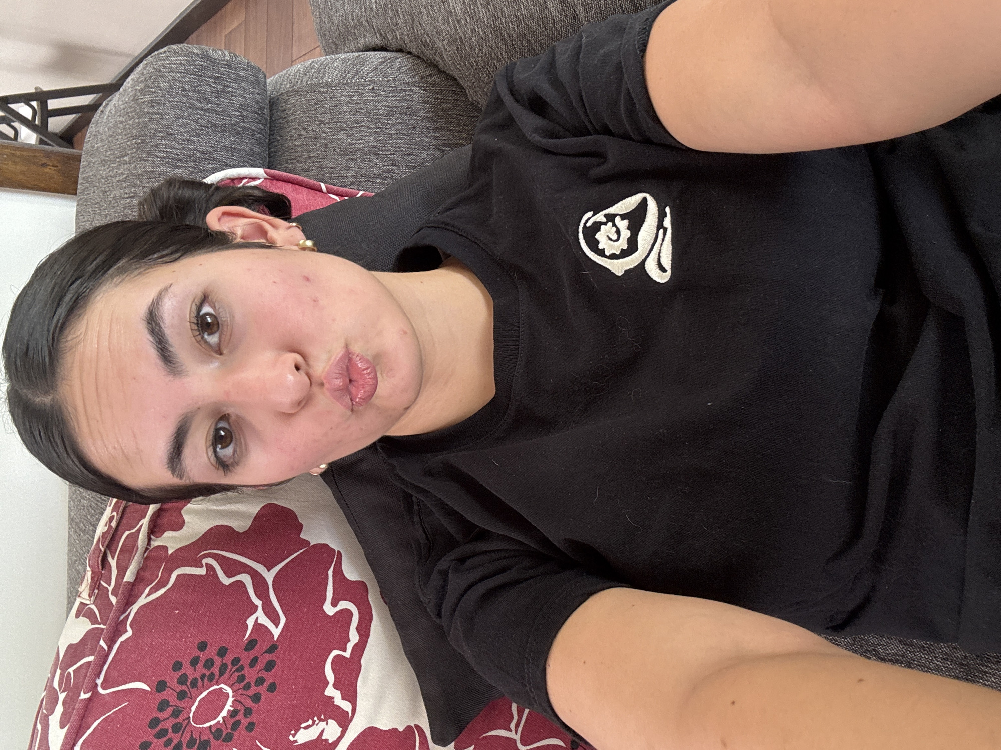 A young woman is shown in a selfie, reclining on a sofa in an indoor setting in San Bernardino Tlaxcalancingo, Mexico. She has dark hair pulled back and is wearing a black t-shirt with a stylized white embroidered logo on the left chest, which appears to be a complex symbol or design incorporating a floral element. A golden stud earring is visible in her right ear. She looks directly at the camera with a playful expression, her lips formed into a pout. Her skin shows some minor imperfections.
She is resting her head and upper body on a decorative pillow featuring a bold red and cream floral pattern, possibly poppies, with black centers. The sofa beneath her is upholstered in a textured, dark gray fabric. In the background, hints of wooden furniture or flooring can be seen. The image captures a casual and relaxed moment, likely during the daytime.