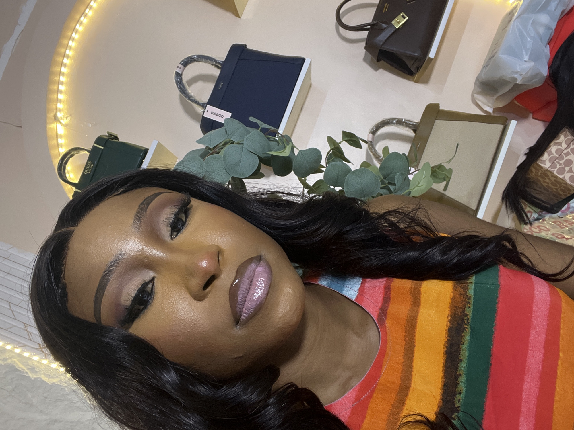 The image is a medium close-up, selfie-style photograph of a young Black woman with long, dark, wavy hair, taken indoors. She is wearing a brightly colored, striped top with horizontal bands of orange, yellow, green, red, and pink. Her makeup is elaborate, featuring winged eyeliner, sculpted eyebrows, and a shimmery, light pink lip gloss. She is looking directly at the camera.
In the background, the wall is painted a neutral tone and decorated with a string of warm white LED lights, arranged in an arched shape. On shelves mounted on the wall are several handbags. The top shelf holds a dark green vanity case with "GUESS EST. 1981" embossed on it. Below that, a navy blue structured handbag with a single top handle is displayed, a white tag hanging from it. Further down, a light beige and brown structured handbag with top handles is visible. On the right side of the image, a dark brown handbag with a gold clasp and two top handles is on a shelf. The lighting in the room is soft and warm, casting a gentle glow.
The specific location within the city of Abuja, Nigeria, cannot be determined from the visual information provided in the image.
