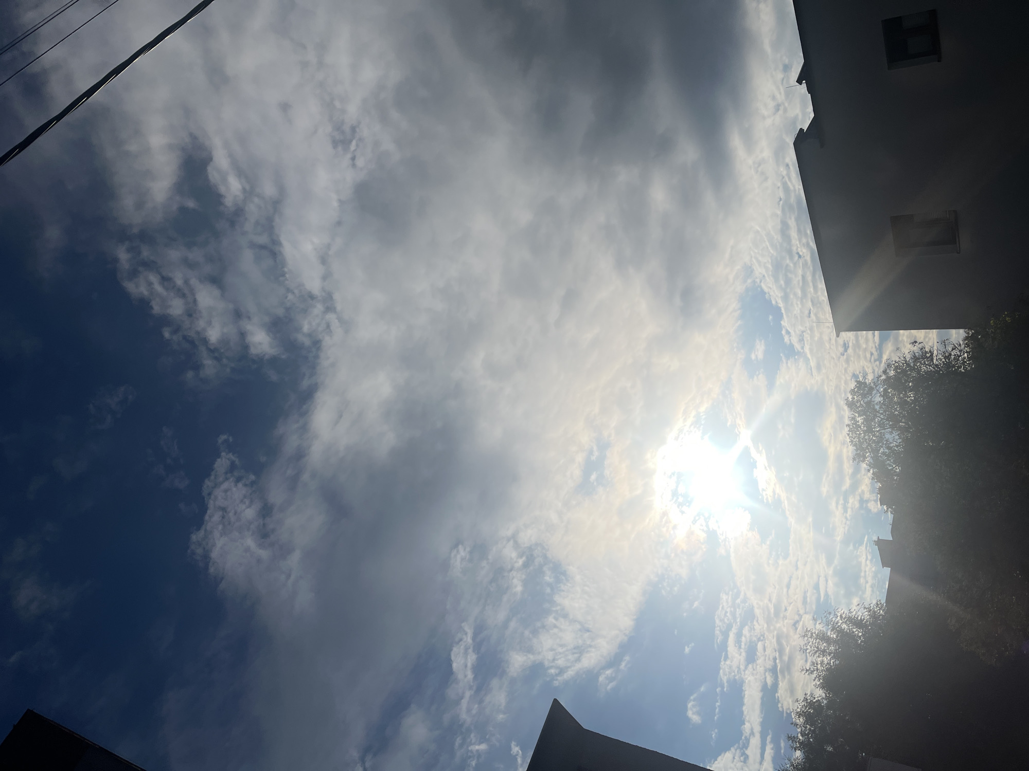 The image depicts a view of the sky from a city. The sky is filled with clouds, with the sun shining brightly through them, creating a lens flare effect. The sun is partially obscured by clouds, which vary in density from wispy cirrus clouds to thicker, darker formations. The deep blue of the sky is visible in some areas. In the foreground and lower portion of the image, the silhouettes of trees and buildings are visible. One building, on the right side, is light-colored and appears to be an apartment building with at least two windows visible. On the left side, the dark silhouette of another building or structure is partially visible. Overhead, power lines are visible, crossing the sky. The overall impression is of an urban landscape with a dynamic sky.