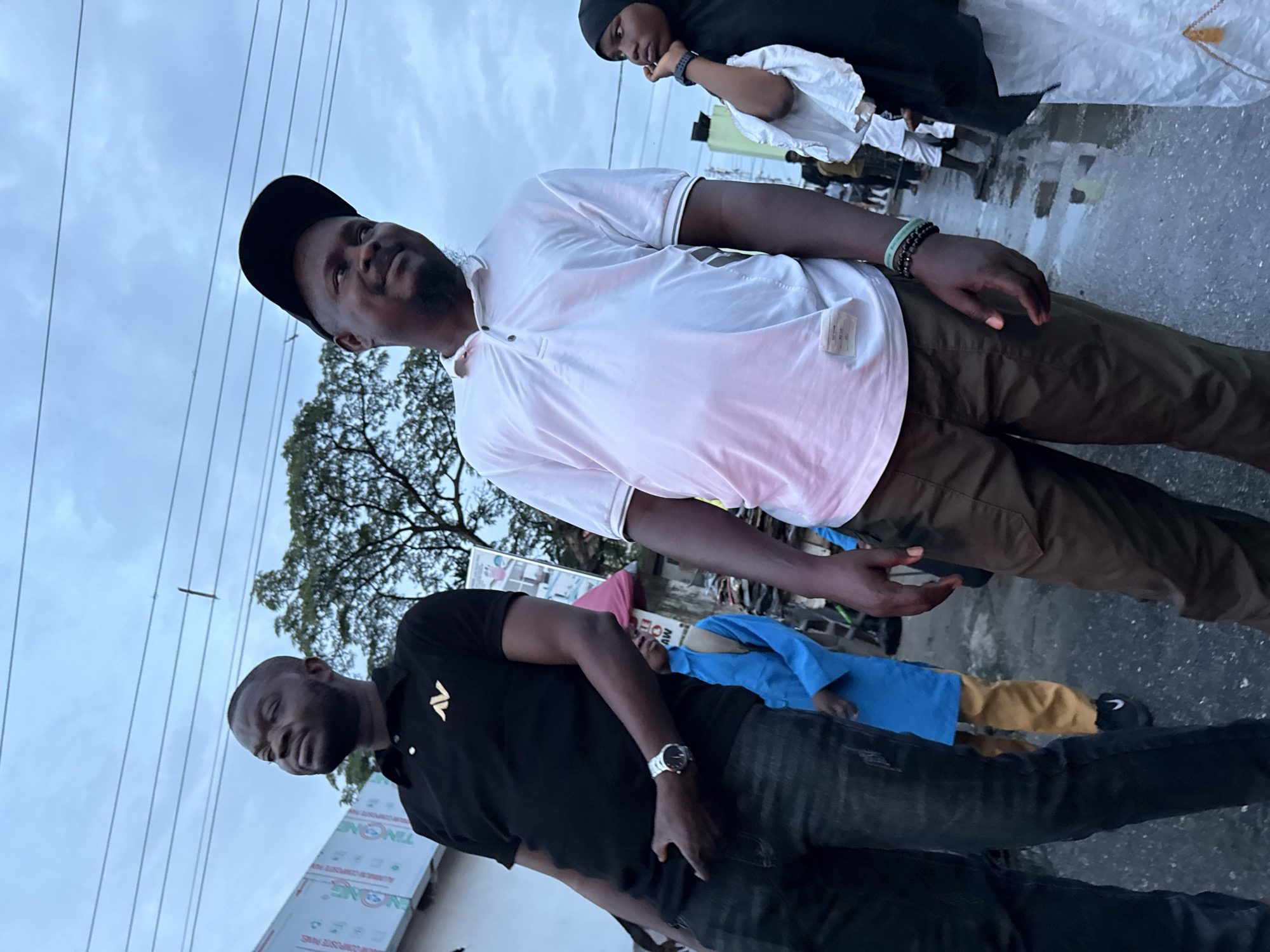 This image was taken on a cloudy day in Ado, Nigeria. In the foreground, two men are looking to the right. The man on the left is wearing a black polo shirt with a gold logo and dark jeans. He has a watch on his left wrist. The man on the right is wearing a white polo shirt and brown pants, with a black baseball cap. He is wearing bracelets on his left wrist. Partially visible on the far right is a woman in a black hijab and white garment. In the middle ground, a person in a blue shirt and yellow pants is walking away from the camera. The background shows a tree and some buildings. The ground appears wet, suggesting recent rain.