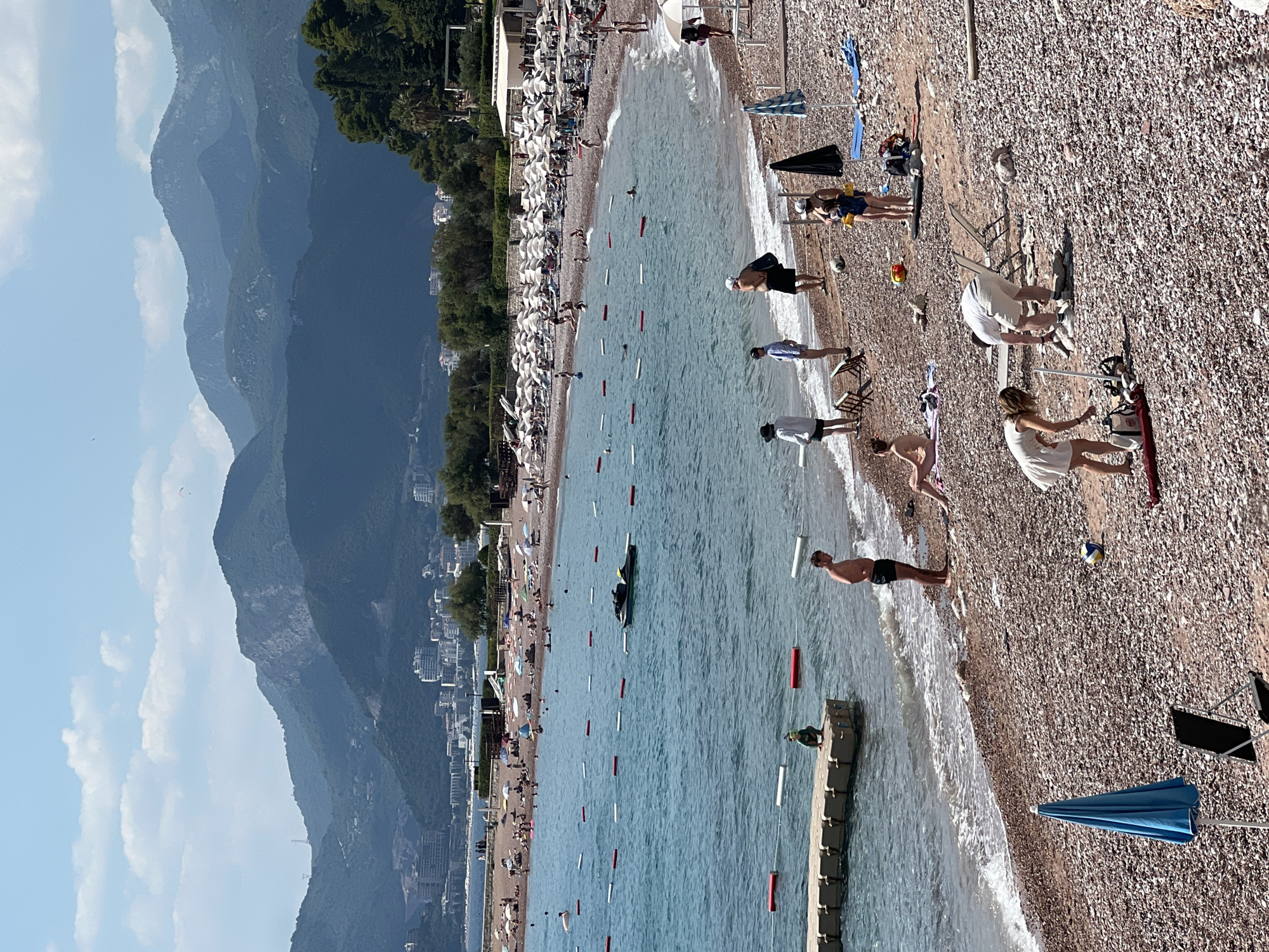 This media file depicts a sun-drenched beach scene in Sveti Stefan, Montenegro. The primary subjects are numerous people enjoying a leisurely day at the coast. They are engaged in various beach activities such as sunbathing, swimming, and walking along the shore. Scattered across the pebble beach are beach chairs, umbrellas, and a volleyball, indicating a recreational setting.
The sea is calm and a clear blue, with gentle waves lapping at the shore. In the background, lush green trees and the distinctive architecture of coastal buildings suggest a populated area. Looming behind the developed coastline are imposing, layered mountains, shrouded in a soft, hazy light, hinting at a clear, warm day. A few clouds dot the bright blue sky, adding texture to the vast expanse.
The overall atmosphere is one of relaxation and vacation. The bright sunlight and the activity on the beach point to it being midday or early afternoon. The clarity of the water and the mountains, despite their distance, suggests good visibility and pleasant weather conditions. The image captures the quintessential experience of a summer day at a European coastal resort, with the dramatic mountainous backdrop adding a unique geographical element. No visible text is present in the image.