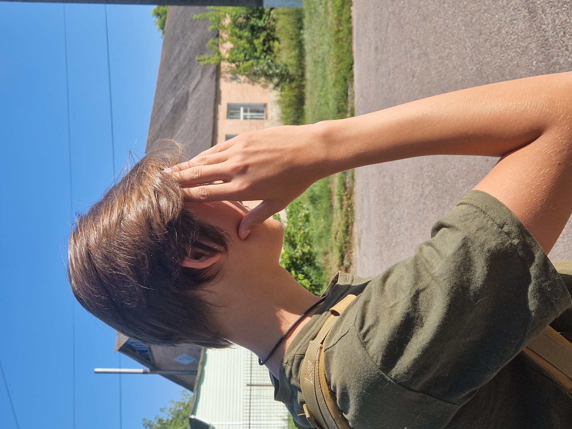 A young person with brown hair is shown from the shoulders up, in profile, with their right hand covering part of their face. They are wearing a green t-shirt and a khaki backpack strap is visible over their left shoulder. They are outdoors, with a paved surface in the foreground and blurred buildings and vegetation in the background under a clear blue sky.