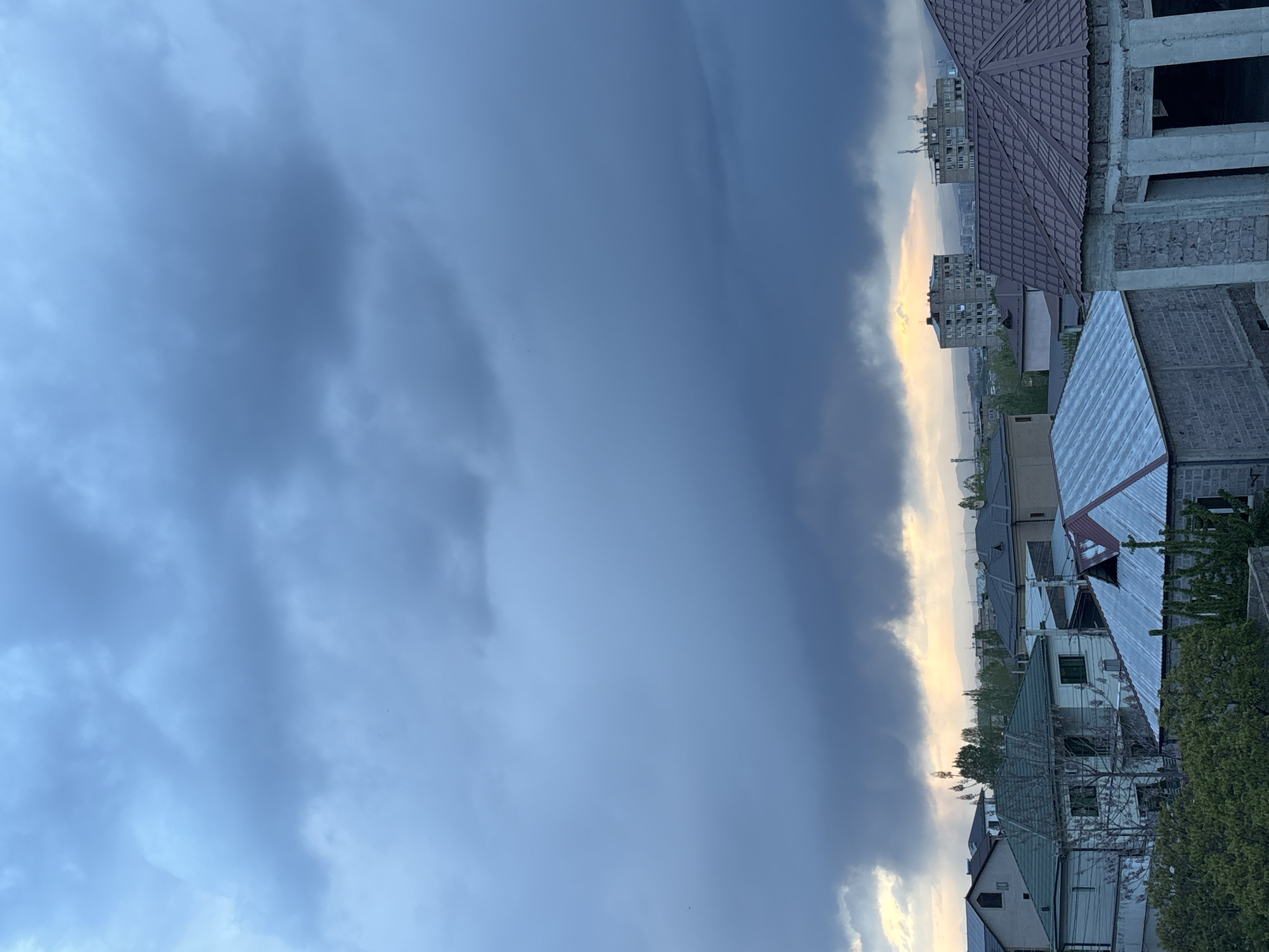 The image depicts a cityscape in Arinj, Armenia, viewed under a cloudy sky with a glimpse of sunlight on the horizon. The foreground shows rooftops of residential buildings, including a partially constructed structure with exposed brick and concrete. In the background, taller apartment buildings are visible. The sky is predominantly overcast with dark, heavy clouds.