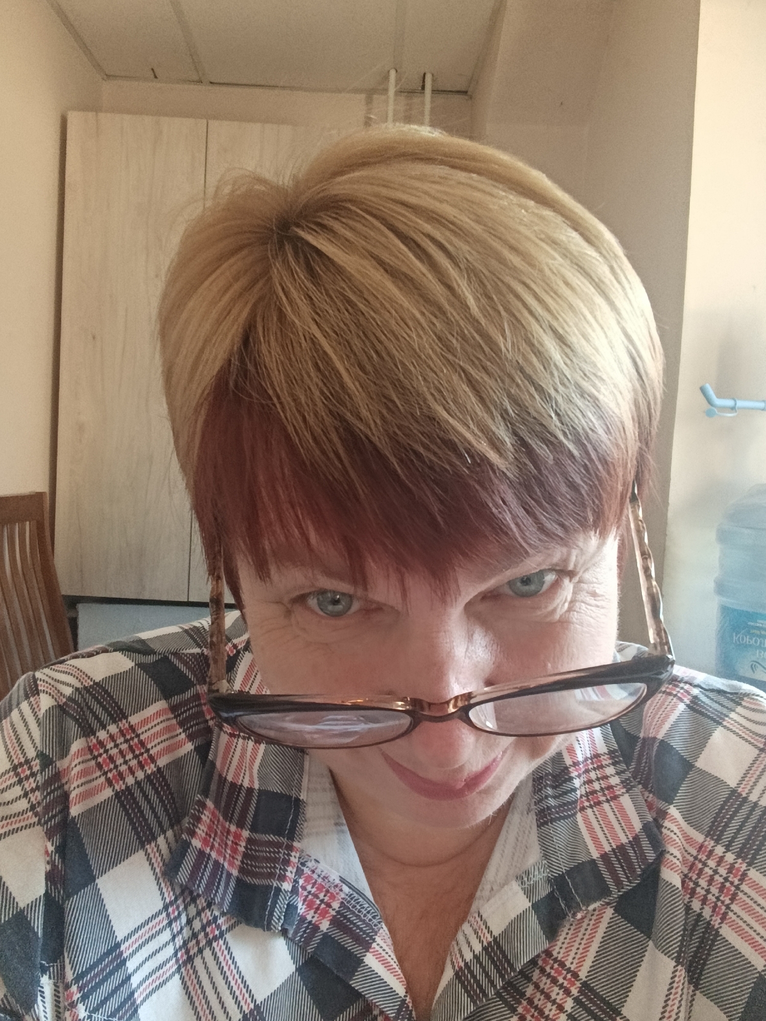 A medium shot captures a woman wearing glasses and a plaid shirt, looking directly at the camera with a slightly lowered gaze. Her hair is styled in a short, blonde cut with reddish undertones near the front. She appears to be indoors, with a light-colored cabinet or wardrobe visible in the background to her left. To her right, a large blue water cooler is partially visible, suggesting a possible kitchen or office environment. The lighting is natural, coming from above. The image does not provide any explicit information to confirm its location in Svietlahorsk, Belarus, as there are no identifiable landmarks or cultural markers.