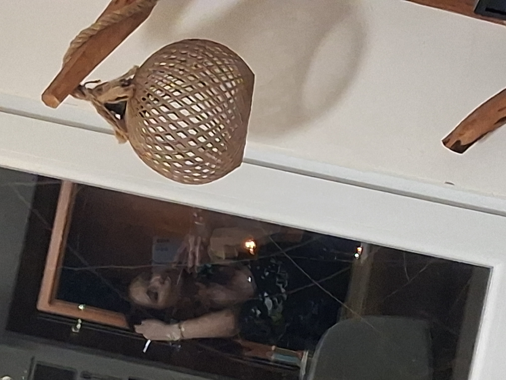 The image shows an interior setting with a woven, globe-shaped pendant lamp hanging from the ceiling. A window reflects a person with long hair, wearing a dark outfit, holding a light-colored object (possibly a cigarette). The location is Unknown, Unknown.