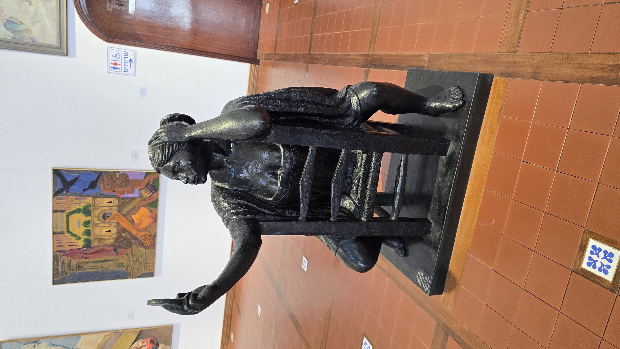 The image shows a dark bronze sculpture of a woman in a squatting position, holding up a smartphone to take a selfie. The sculpture is placed on a black rectangular base on a terra cotta tiled floor. The woman is wearing a draped garment and has her hair pulled up. In the background, there are paintings on a white wall. To the right of the sculpture, there is a wooden door and a sign indicating restrooms. The floor has a pattern of alternating square terra cotta tiles and wooden strips. A small decorative tile with a blue and white floral pattern is visible on the floor in the foreground. This sculpture is located in a museum in Hof HaCarmel Regional Council, Israel.