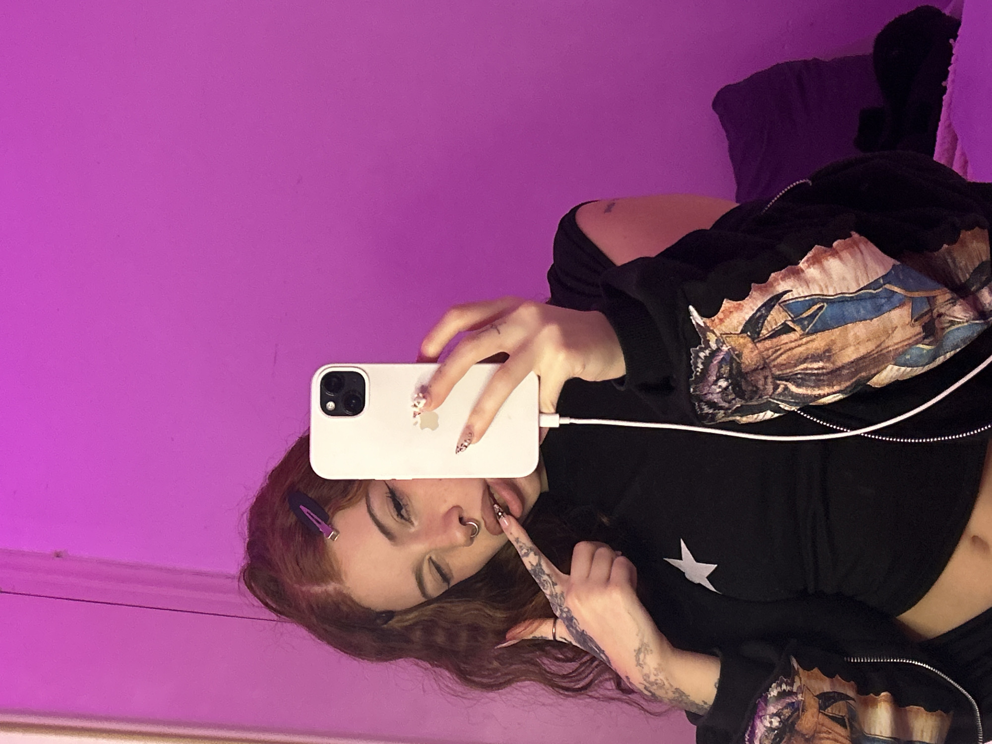 A young woman with light skin and light brown hair poses for a selfie indoors against a solid purple wall. She is lying down, holding a white iPhone in her right hand. She is wearing a black zip-up jacket with a printed design on the sleeves. Her hair is parted to the side, with a purple clip visible. She has a nose ring and her tongue is slightly visible as she touches it with her right index finger. Her right arm and hand are tattooed. The image is brightly lit with a purple hue. There is no indication of the location in the city of Buenos Aires, Argentina.