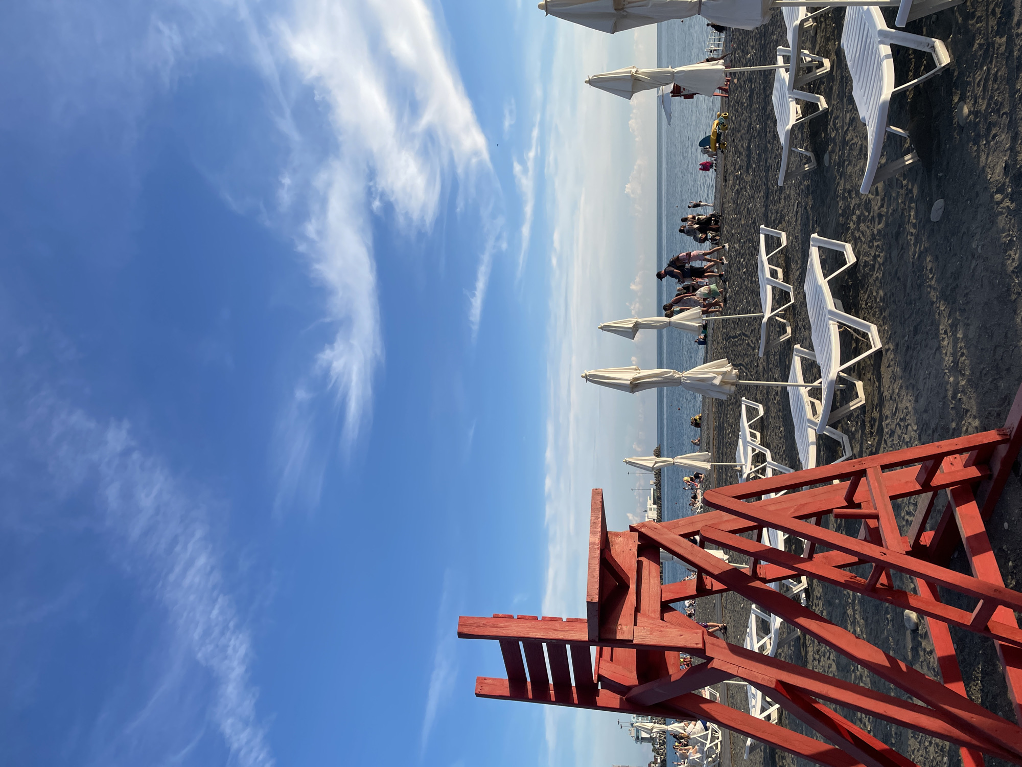 This is a medium-shot, eye-level photograph of a beach in Sochi, Russia, under a bright blue sky with wispy white clouds. In the foreground, a large, weathered red lifeguard chair occupies the left side of the frame, its wooden structure weathered and showing signs of age. Beyond the chair, numerous white lounge chairs are arranged on the dark, pebbly sand, some folded and some open. Several white beach umbrellas are also visible, most closed. In the middle ground, a group of people, appearing to be tourists, are gathered near the water's edge, some walking, some standing. The sea is calm and stretches to the horizon, where a faint line of buildings and possibly a port can be seen in the distance. The overall atmosphere is one of a typical, sunny beach day.