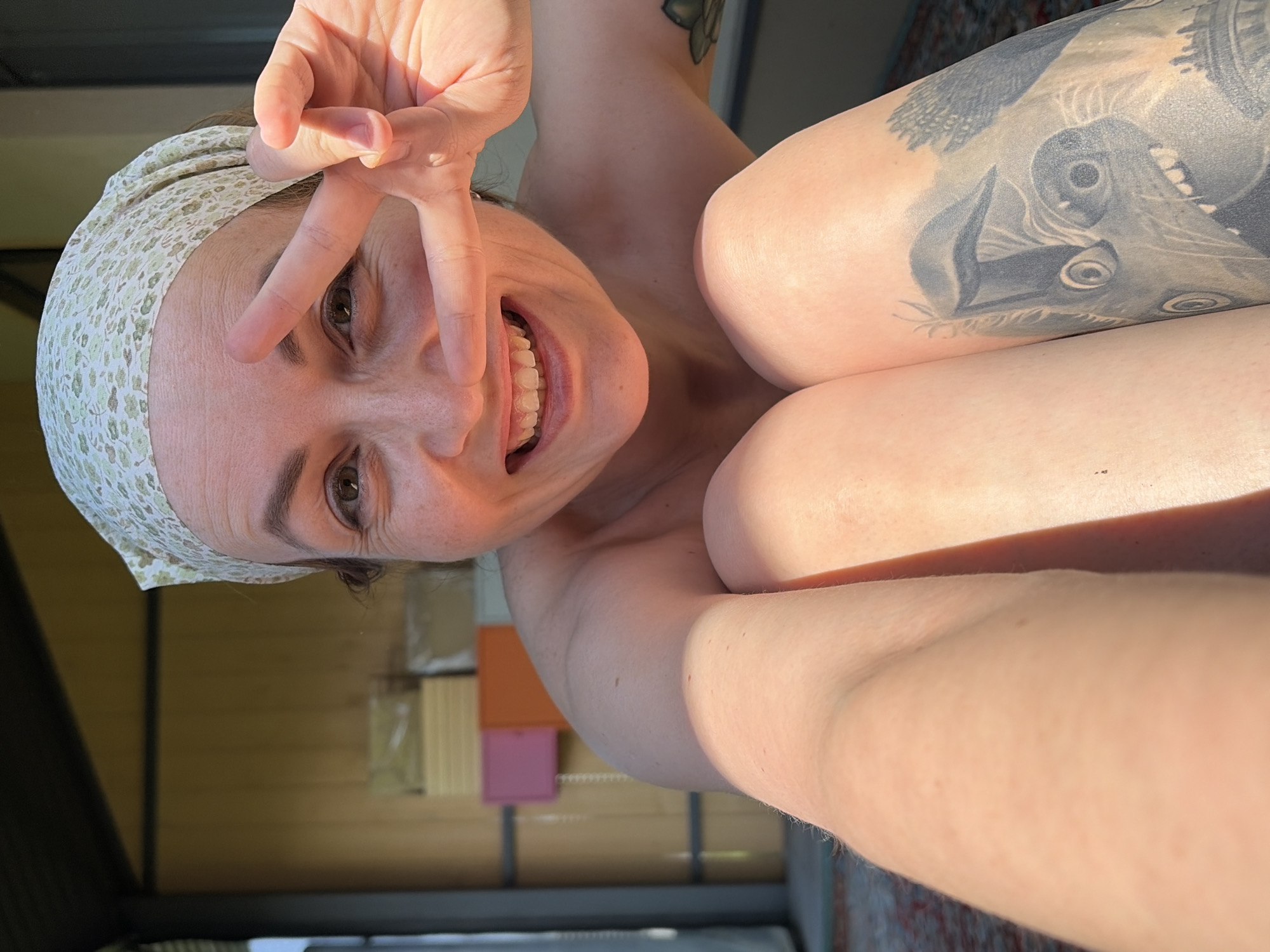 A woman is taking a selfie outdoors. She is smiling and holding up a peace sign with her fingers. She is wearing a headscarf. The location appears to be in Chaikovsky, Russia. The background shows a wooden wall and some stacked boxes. She has a tattoo on her upper arm.
