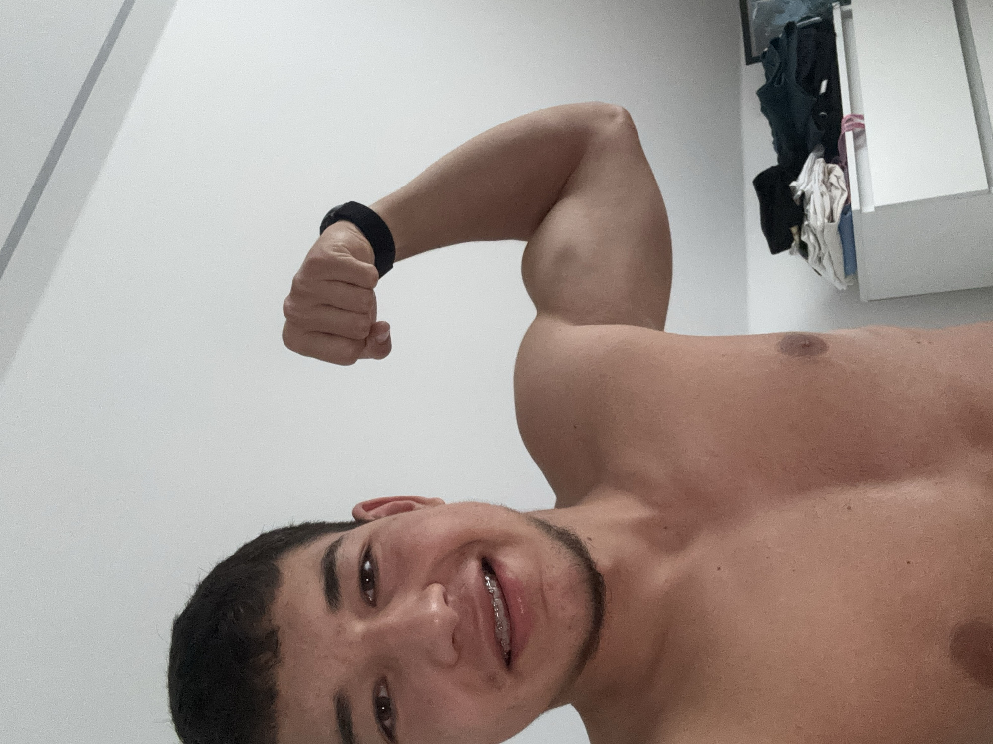 The image shows a young man with short dark hair, brown eyes, and braces on his teeth, flexing his bicep. He is shirtless and has a black fitness tracker on his left wrist. Behind him, a white dresser is visible with clothes stacked on top. The setting appears to be indoors, likely a bedroom, with a white wall in the background. There is no indication of the location in Viernheim, Germany.
