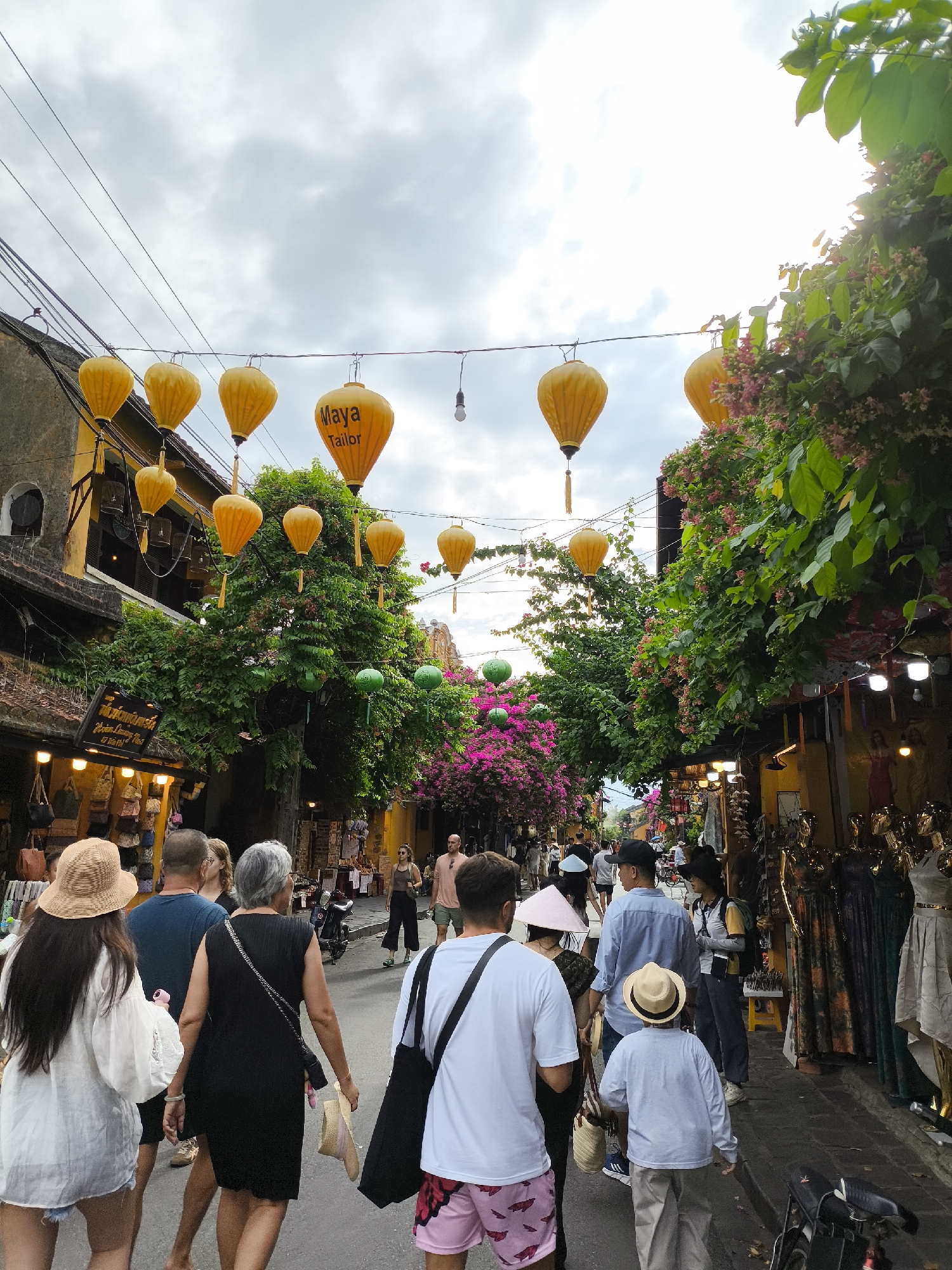 This image depicts a street scene in Hoi An, Vietnam, characterized by traditional yellow lanterns and lush greenery. In the foreground, a group of people are walking away from the camera down a narrow street lined with shops. A woman in a white shirt and straw hat is on the left, followed by a man in a blue shirt, a woman in a black dress, and a man in a white t-shirt and patterned shorts. To the right, a young boy in a straw hat walks ahead of other pedestrians. The shops on either side of the street display merchandise, including clothing on mannequins. Overhead, yellow lanterns hang from wires, some featuring the "Maya Tailor" logo. The architecture is traditional, with yellow facades and balconies. Green trees and pink flowers cascade down the sides of the buildings, adding to the vibrant atmosphere. The sky is overcast, suggesting either early morning or late afternoon.