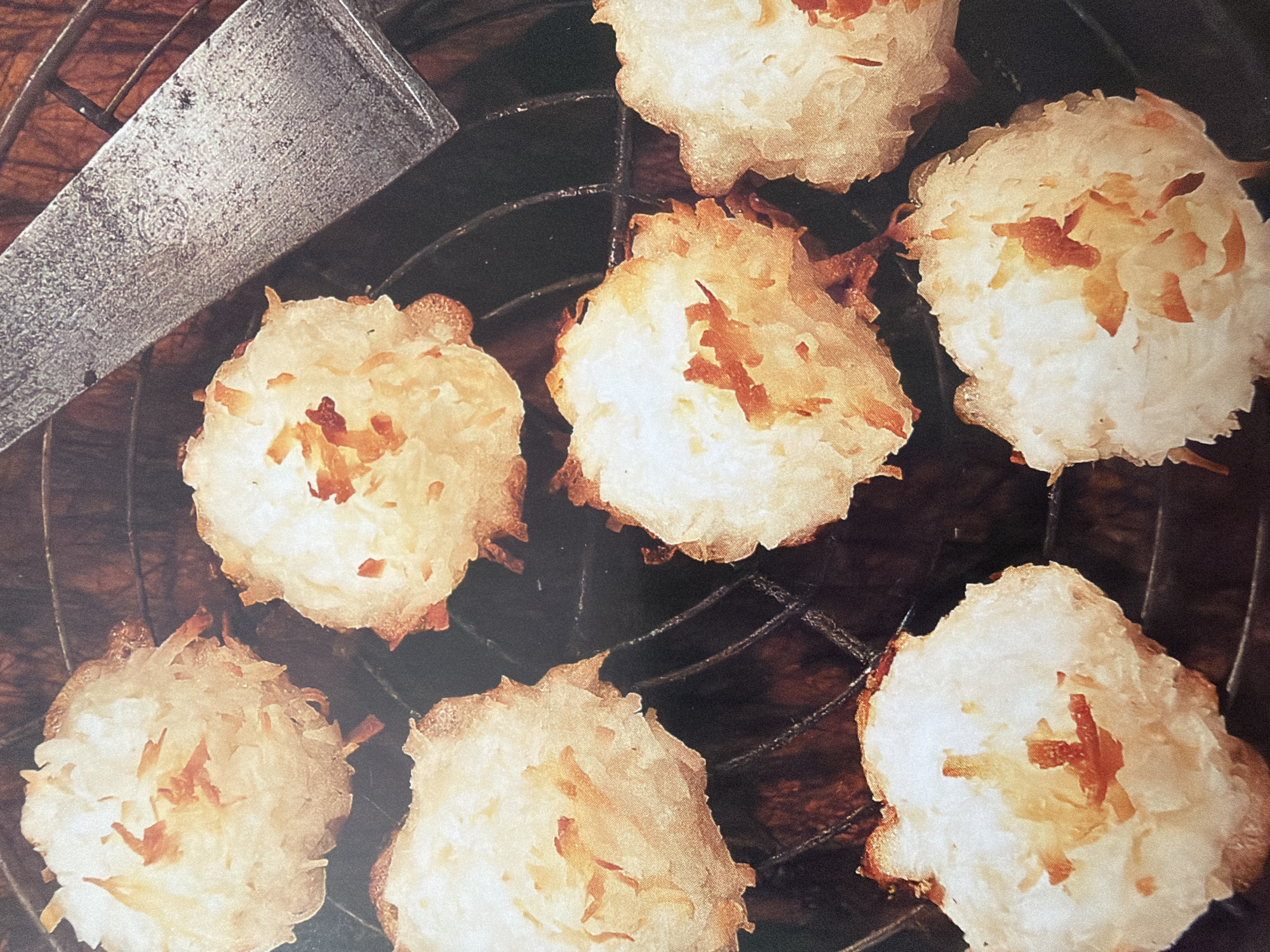 A close-up overhead shot shows seven golden-brown coconut cookies arranged on a dark metal cooling rack. The cookies are roughly spherical and have a textured surface with visible shreds of coconut and some darker, caramelized bits. A dull silver metal spatula with a textured handle rests diagonally across the upper right portion of the frame, partially obscuring some of the cookies. The background is dark and appears to be a wooden surface.