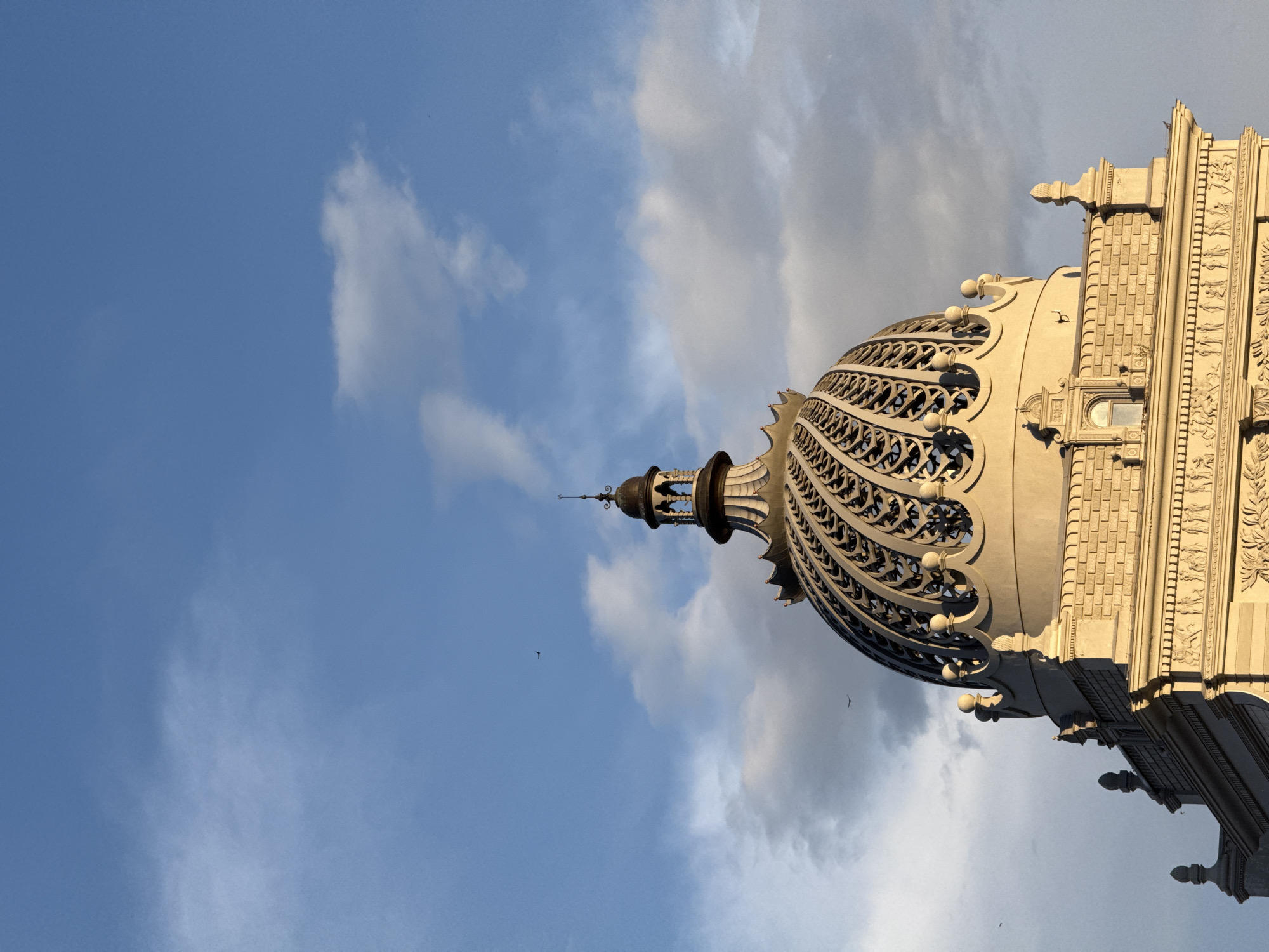 The image shows the dome of the Kazan State Circus, in Kazan, Russia. The dome is ornate and appears to be constructed of metal and light-colored stone. The sky behind the dome is light blue with white clouds. There are at least three birds visible in the sky. The image is oriented vertically, showing the dome rising into the sky.