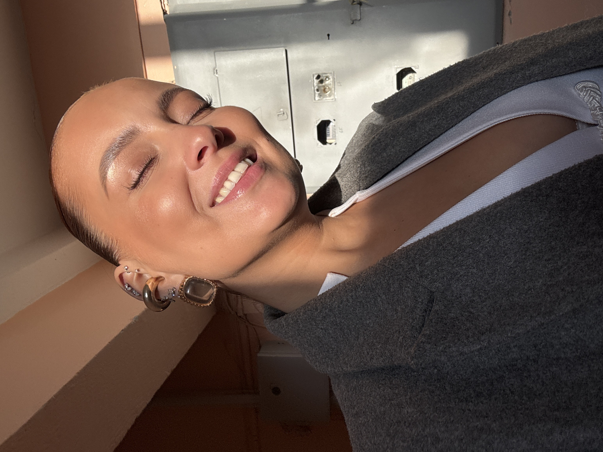 A close-up photograph captures a female subject from the chest up, positioned in what appears to be an urban outdoor or semi-outdoor setting. The subject, with pulled-back dark hair, has her eyes closed and is smiling broadly, revealing white teeth and pink lips. Her skin exhibits highlights consistent with strong direct sunlight, particularly on her forehead, cheekbones, and nose.
She wears a light-colored, possibly white, V-neck top, partially visible beneath a dark gray, textured coat or jacket draped over her shoulders. The visible left ear is adorned with multiple pieces of jewelry: several small, clear stud earrings are placed along the upper helix, a larger golden C-shaped earring or cuff is visible on the mid-helix, and a golden drop earring with a dark, rectangular stone hangs from the lobe.
The background consists of architectural elements. To the upper left, a light brown/beige, possibly concrete or painted, ceiling or overhang is visible, casting a shadow. To the right and behind the subject, a large, utilitarian gray panel or wall is present. This panel features several recessed cutouts, some appearing empty, others containing unclear components or debris, indicative of an industrial or functional structure.
The scene is illuminated by strong, directional sunlight, casting clear shadows and creating high contrast. The background elements are generic and do not provide specific markers to identify a precise location within the city of Chaikovsky, Russia.