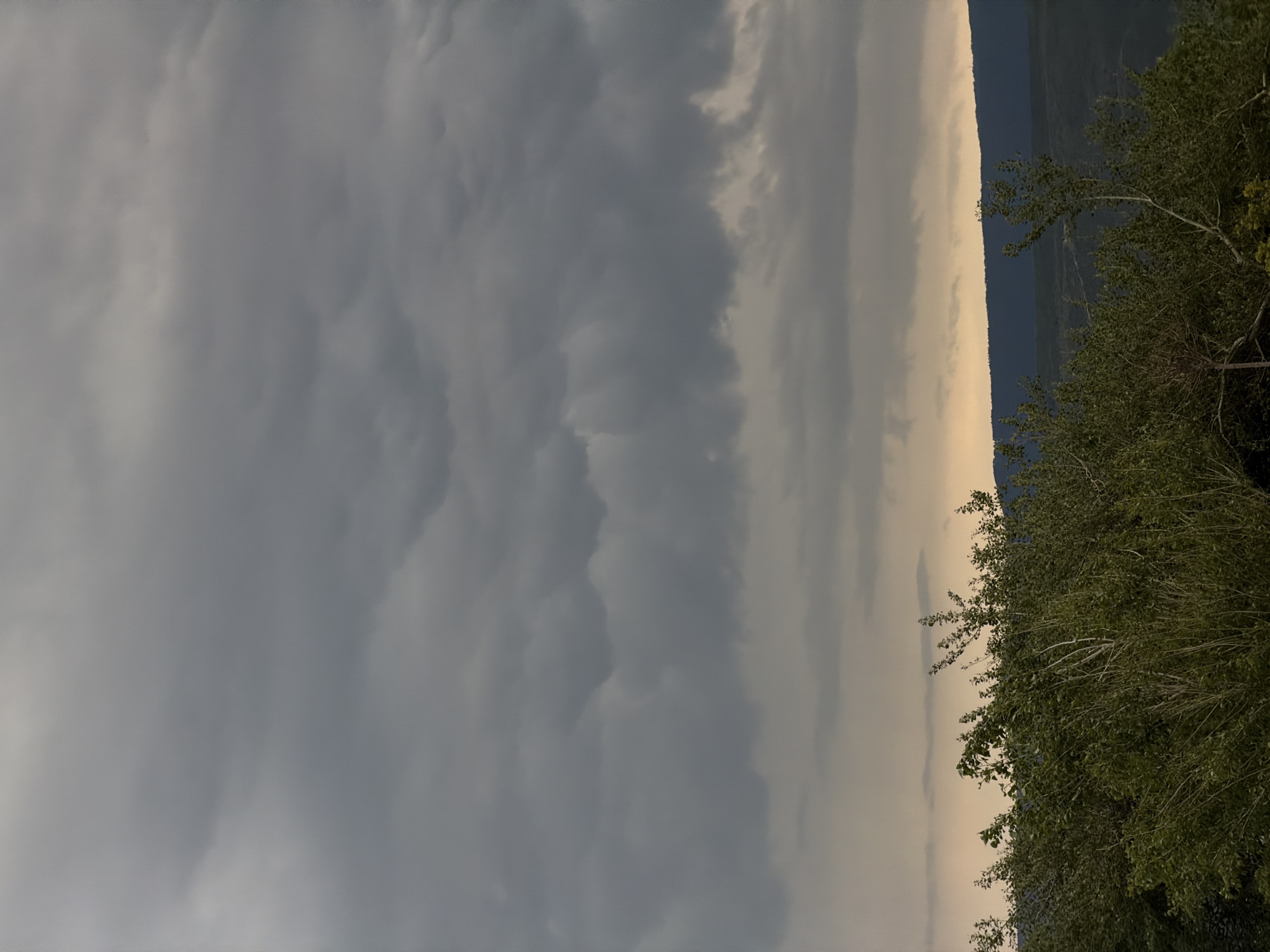 The image shows a landscape with a heavy, overcast sky filling the left side. The right side of the image features a dark, hilly or mountainous terrain, partially obscured by trees. The trees are dense with green leaves. A band of light, possibly from the sun trying to break through the clouds, separates the dark mountains and trees from the heavy sky. The location is Mukhatgverdi, Georgia.