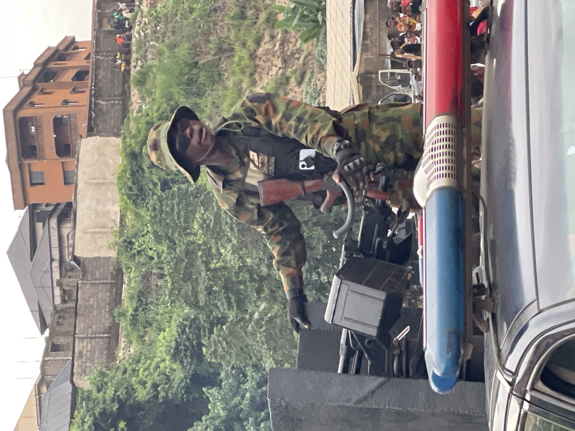 A soldier in camouflage uniform, a wide-brimmed hat, and sunglasses is standing on top of a dark-colored vehicle. The soldier is holding a rifle and appears to be looking to their right. The vehicle has a red and blue light bar mounted on its roof. In the background, there are buildings, trees, and a group of people watching. The scene suggests a public setting with a uniformed presence, possibly for security or a parade.