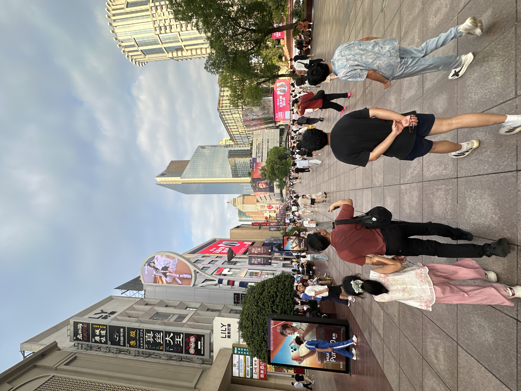 The image depicts a bustling pedestrian street in Shanghai, China, likely in a commercial district like Nanjing Road or Huaihai Road, characterized by tall buildings and an abundance of shops and advertising. Several individuals are walking along the wide, paved walkway. In the foreground, from left to right, are a young girl in a white sleeveless top with pink floral embroidery and loose pink pants, holding hands with a man in a dark red polo shirt and black pants. Behind them, two young men are walking away from the camera; one in a black t-shirt and dark shorts, the other in a light blue patterned shirt and matching pants.
The environment is urban and commercial. Buildings line both sides of the street, featuring large illuminated signs and advertisements. Prominent signage includes "LILY 商务时装" (LILY Business Fashion) and a large digital billboard with an image of a woman. In the background, modern skyscrapers with reflective glass facades punctuate the skyline under a cloudy sky. Trees are visible along the sides of the pedestrian area, providing some greenery amidst the urban landscape. The overall scene conveys a sense of everyday city life and commercial activity in Shanghai.