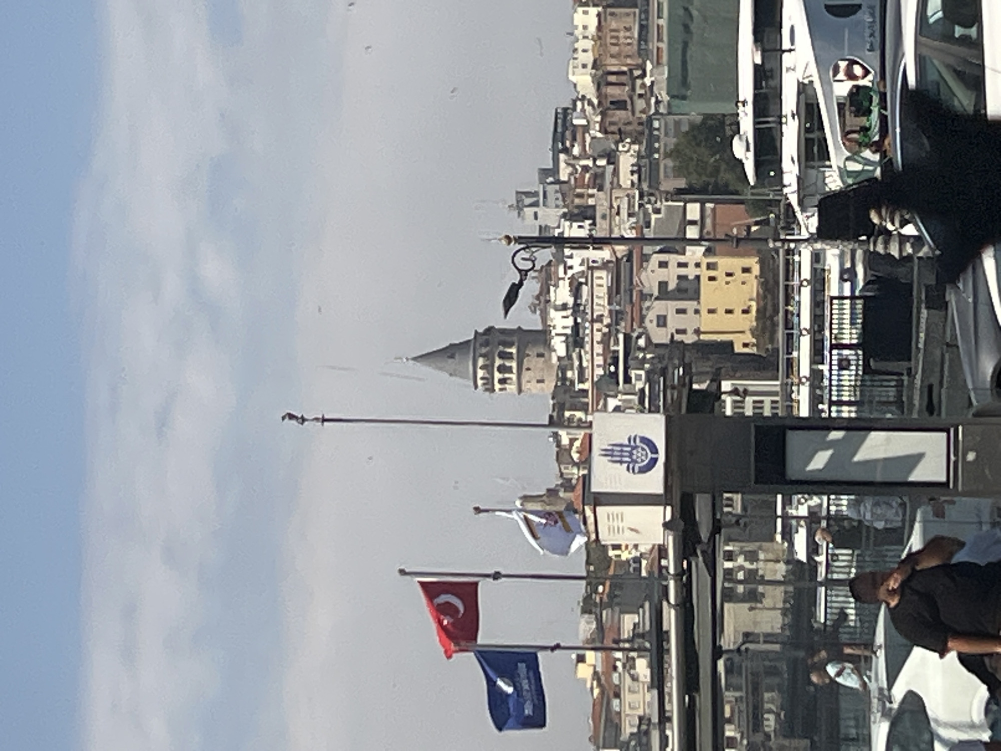 The image shows a view of the Galata Tower in Istanbul, Turkey, from across the water, likely the Golden Horn. In the foreground, there are several flags, including the Turkish flag and a blue flag with white writing. A large, gray structure with a white panel displaying a blue hand symbol is also visible. Several boats are docked or moving in the water. In the background, the Galata Tower is prominently featured, surrounded by a densely populated cityscape with buildings of varying sizes and colors. The sky is partly cloudy. Several people are visible in the foreground, near vehicles and what appears to be a bus stop or ferry terminal.